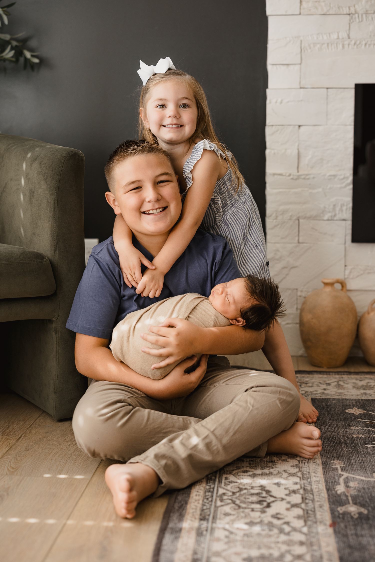 A smiling boy sits cross-legged holding a newborn while his little sister with a bow hugs him from behind indoors.