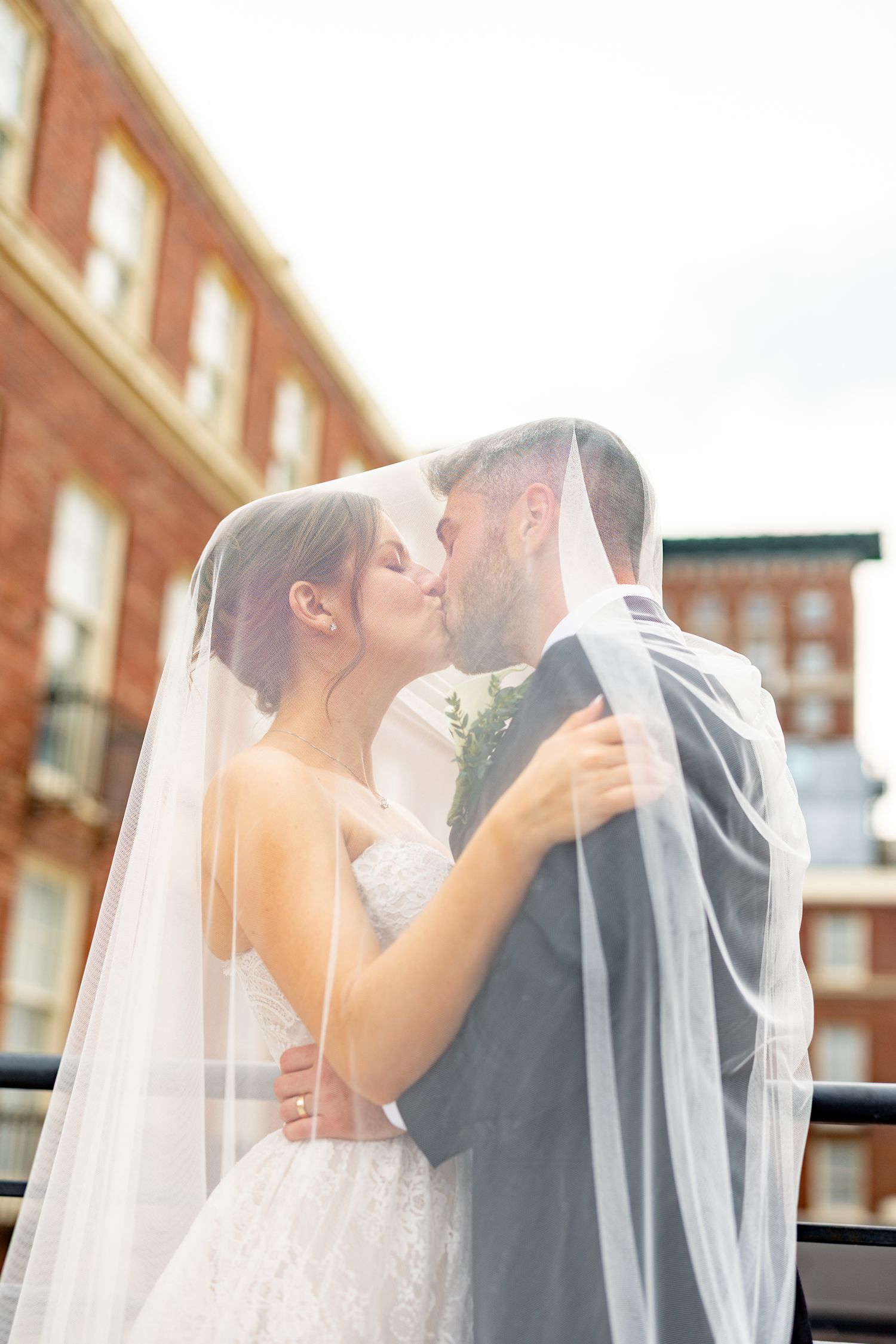 Bride and groom share a romantic kiss under a flowing white veil with brick buildings in the background.