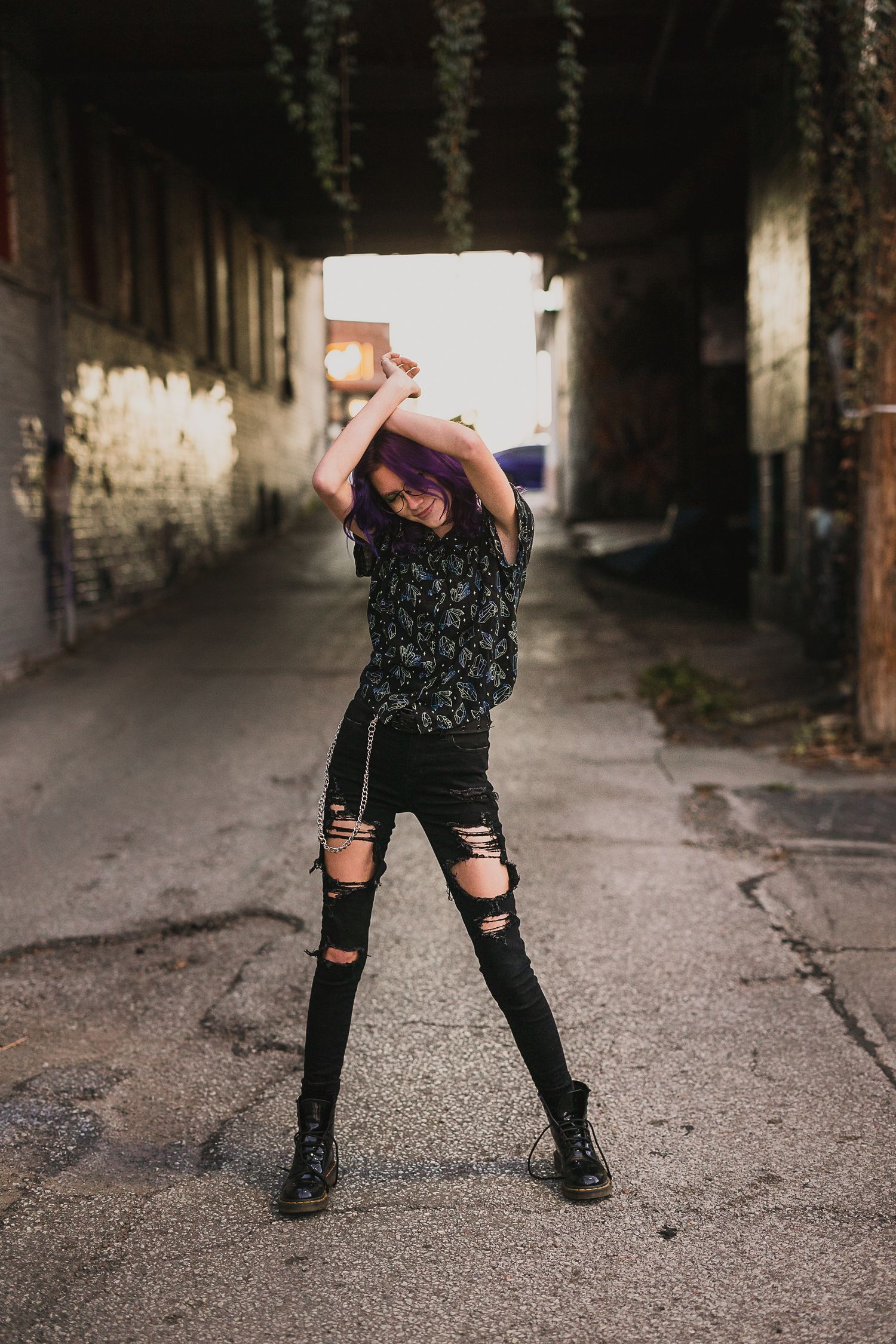 Young woman with purple hair in ripped black jeans and boots poses in an urban alley with hanging vines.