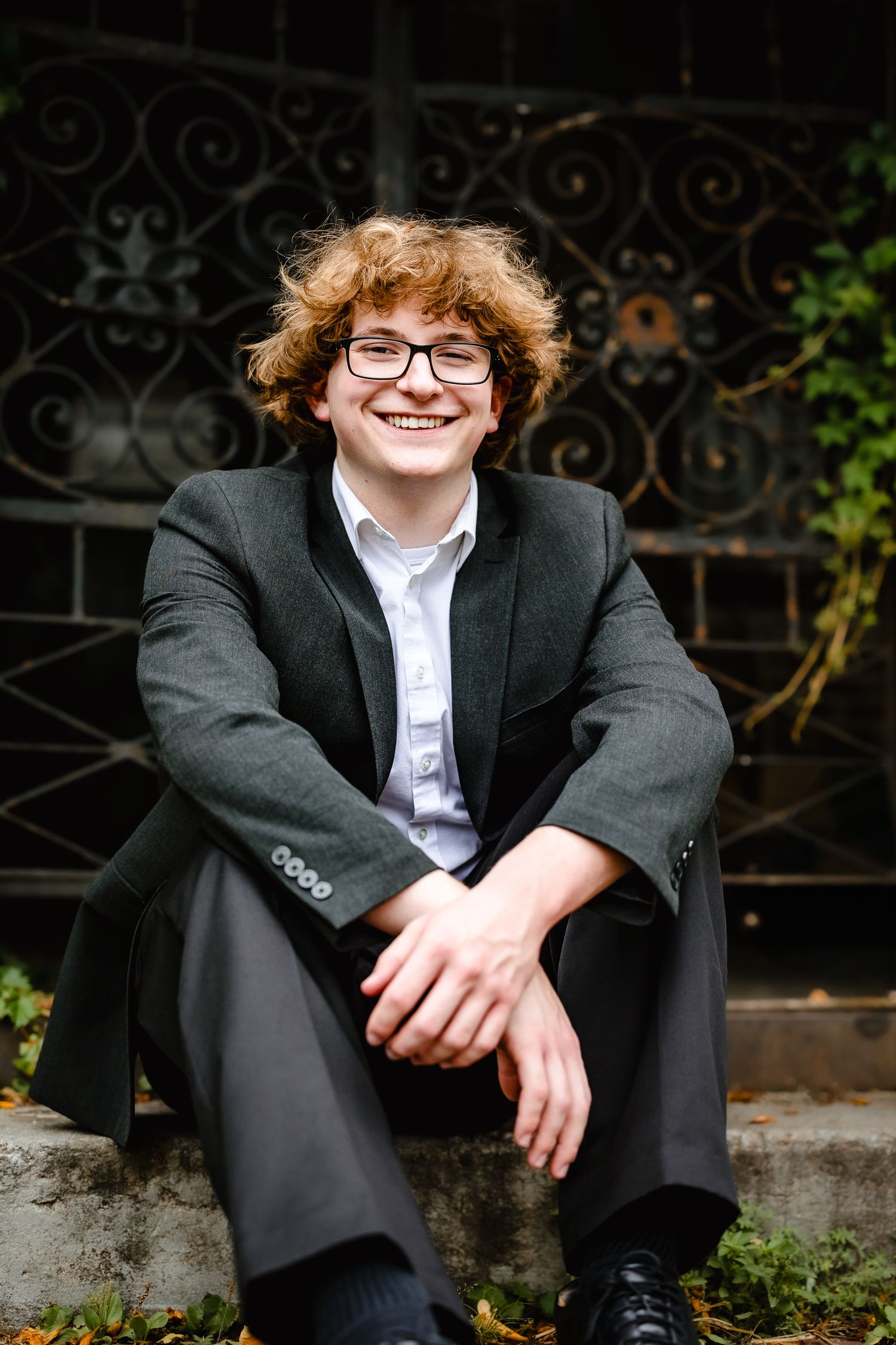 Smiling teenage boy with curly red hair and glasses sits on steps wearing a dark blazer and white shirt.