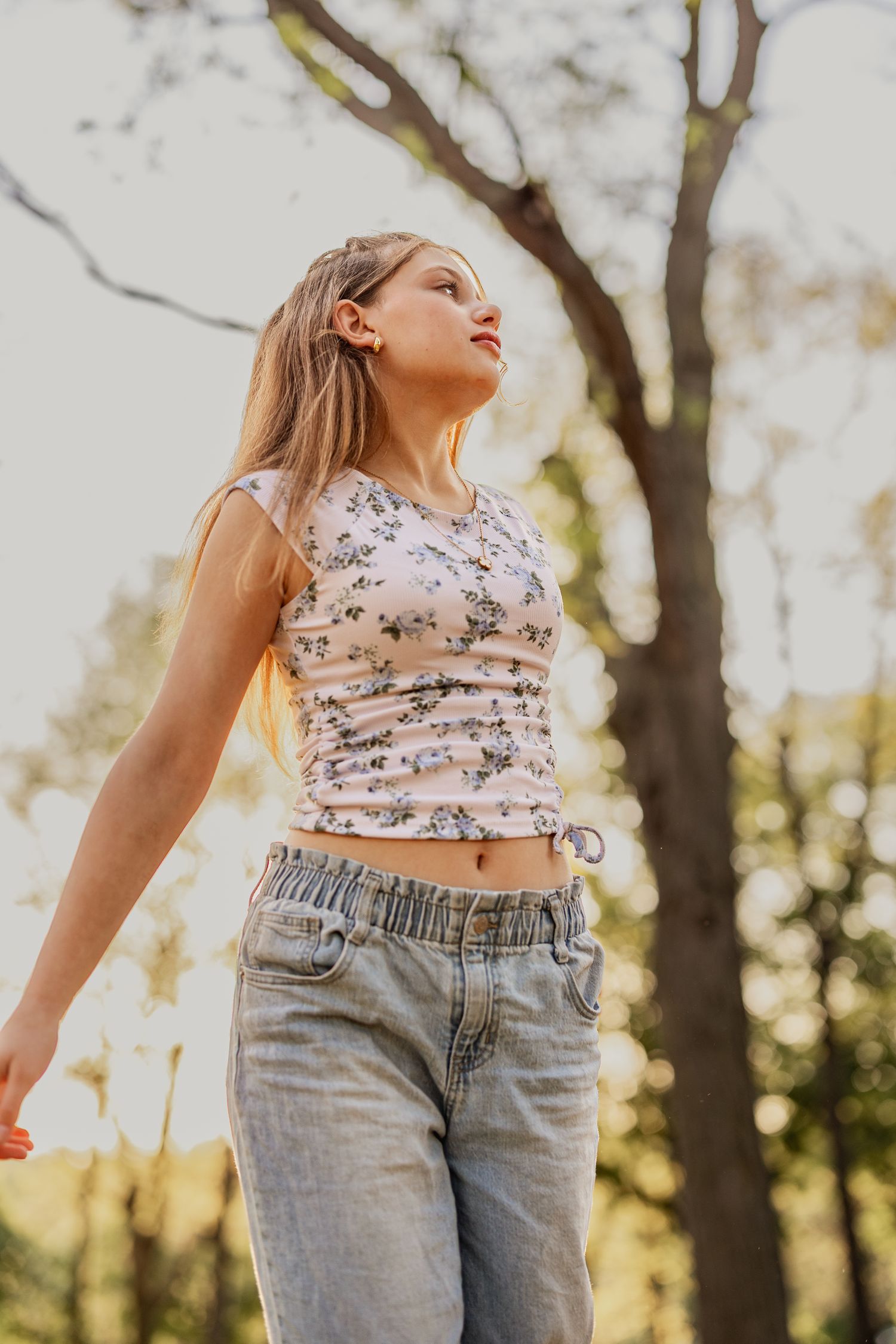 Young woman in floral crop top and jeans standing outdoors in sunlit park with trees in background.