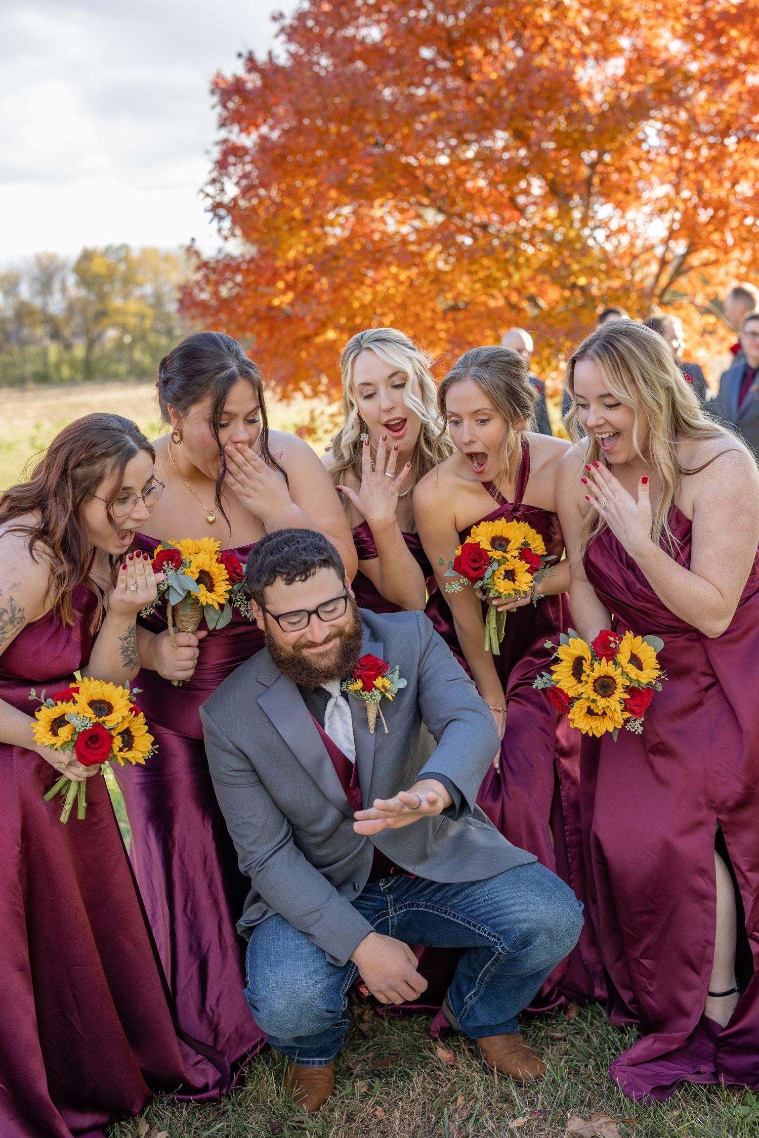 Groom kneels showing ring to surprised bridesmaids in burgundy dresses, holding sunflower bouquets, with vibrant fall foliage backdrop.