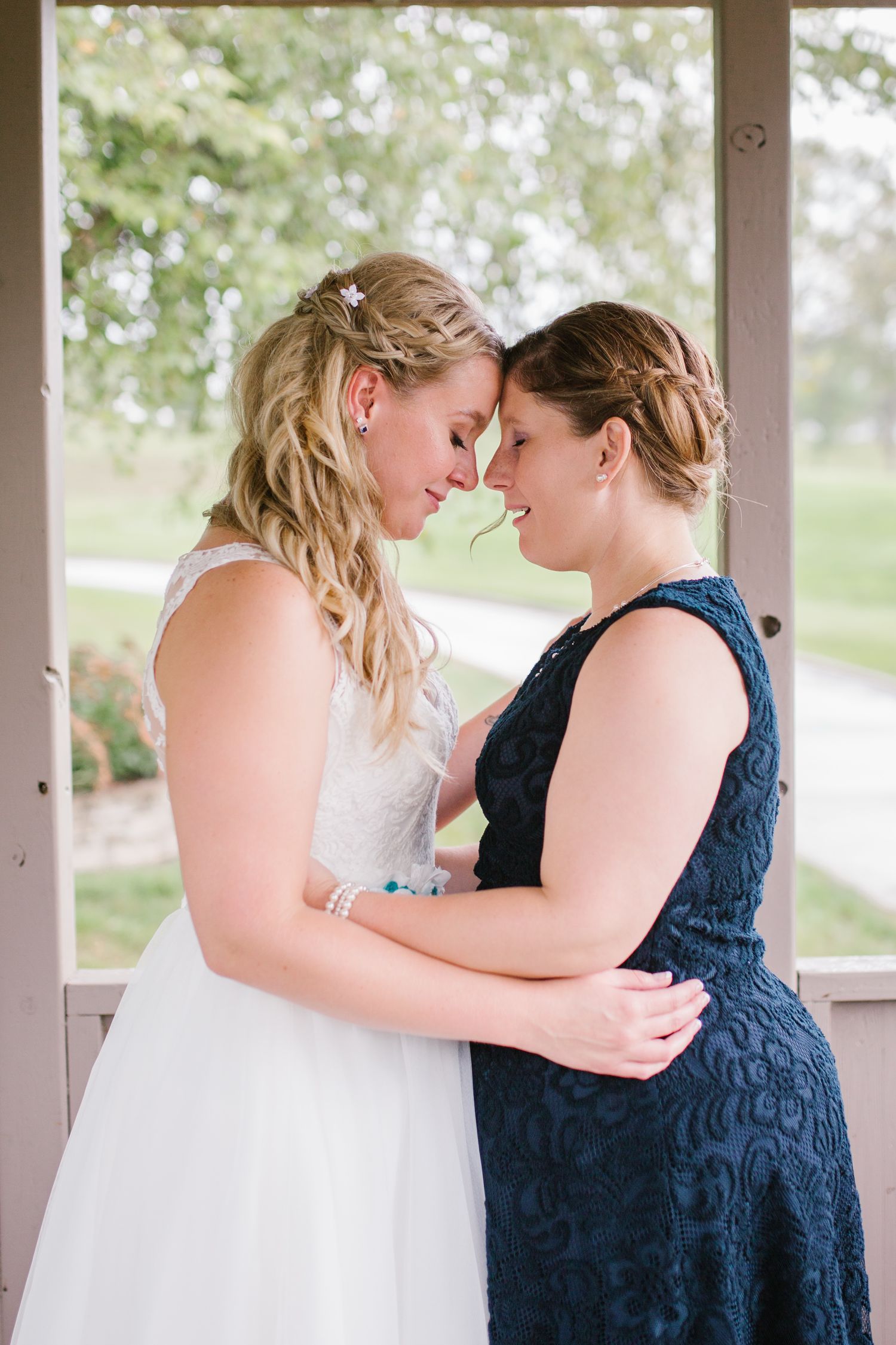Two brides share an intimate forehead touch moment, one in white lace gown and one in navy blue dress, at their wedding.
