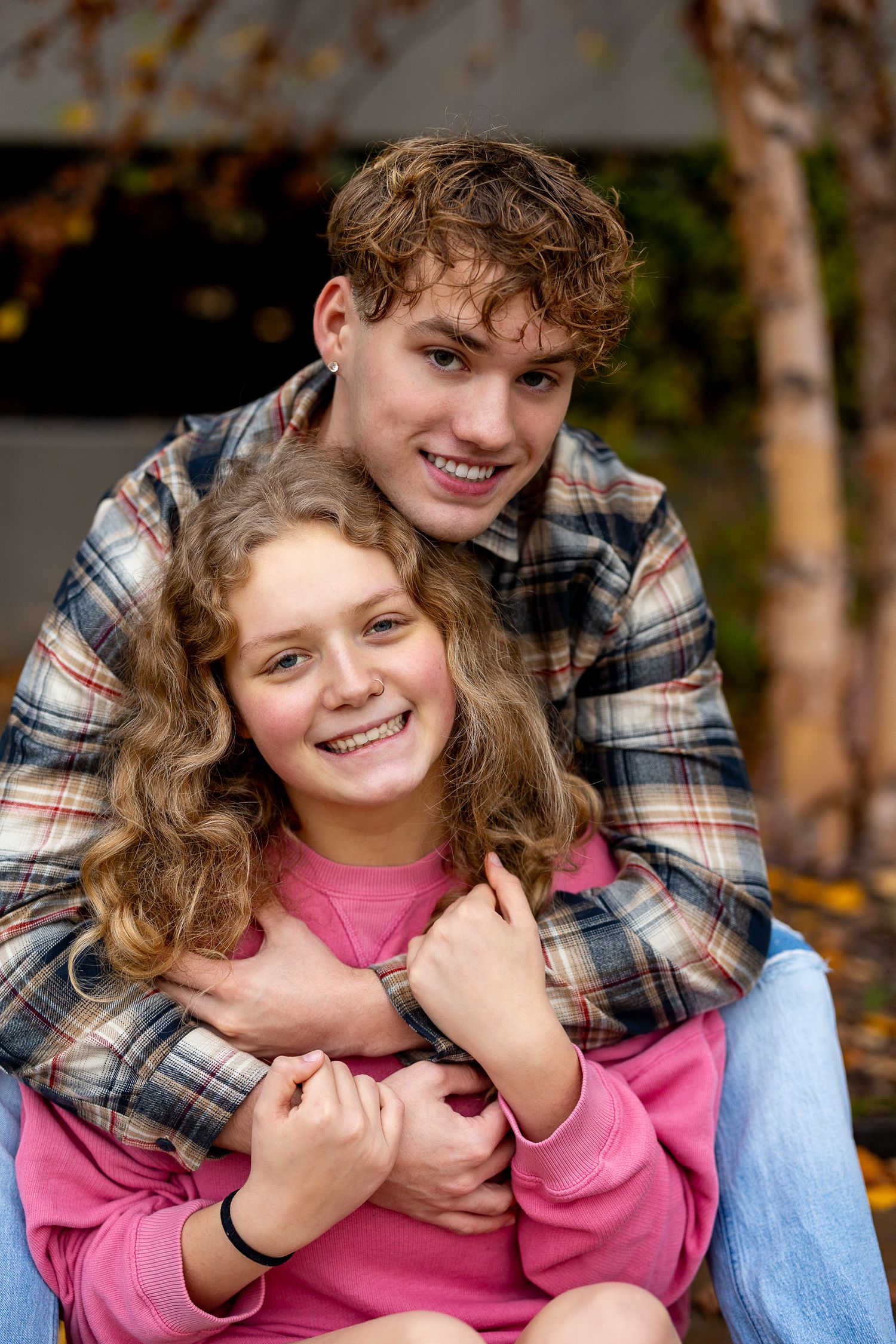Two smiling teenagers embrace outdoors in autumn, boy in plaid shirt hugging girl in pink sweatshirt.