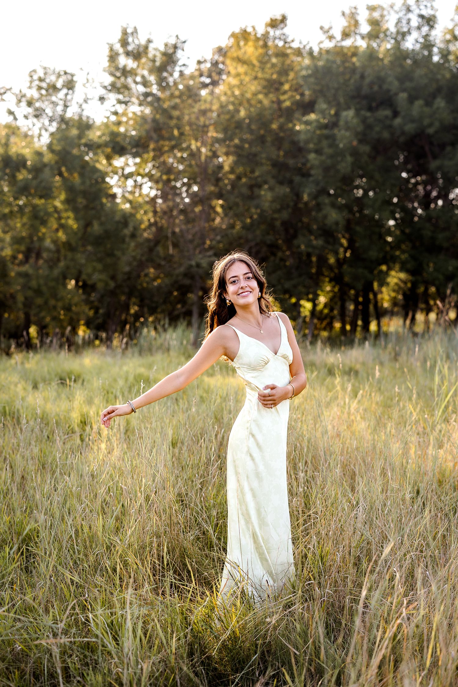 Smiling young woman in white satin slip dress standing in golden meadow with trees at sunset.