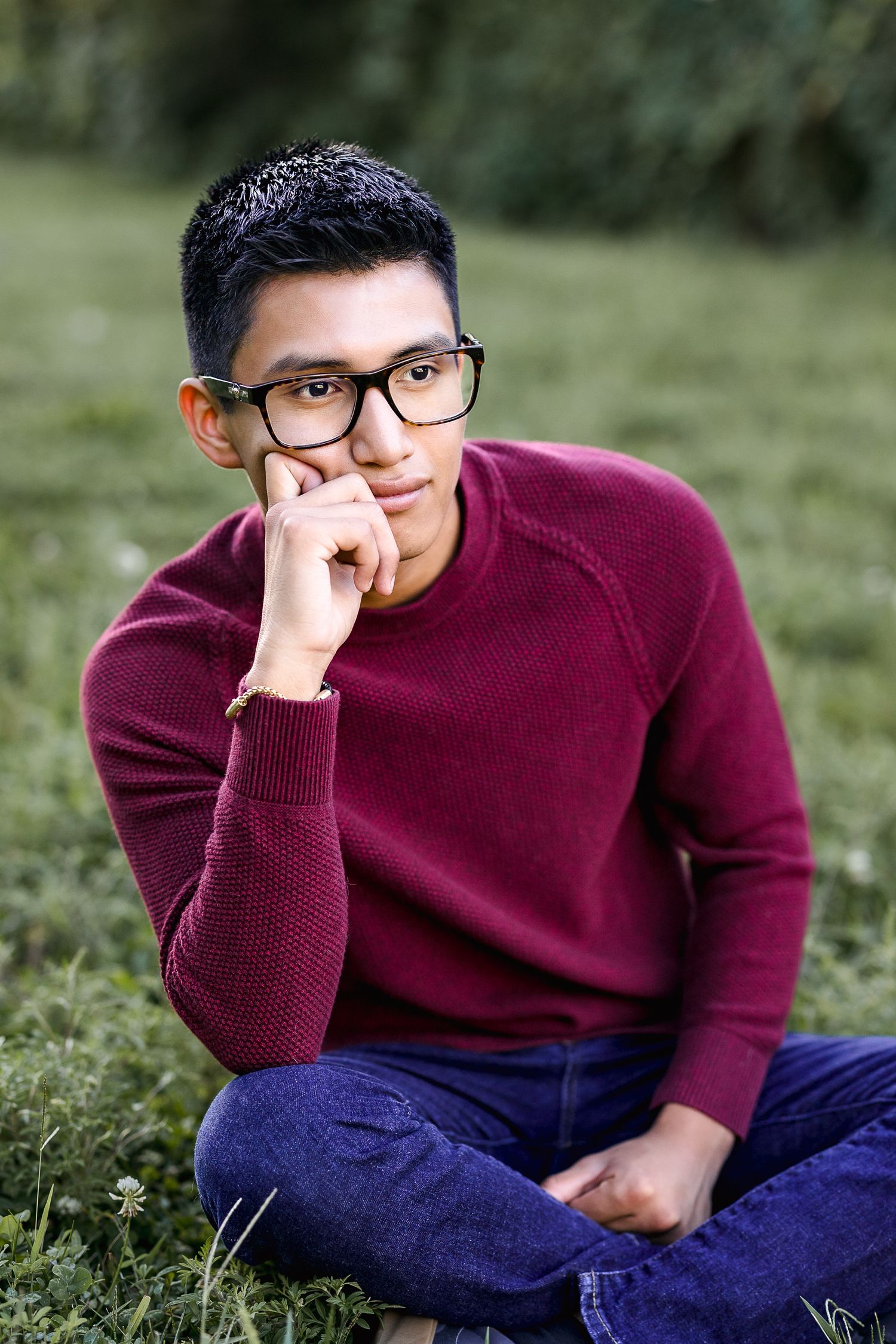 Young man in red sweater and glasses sitting cross-legged on grass, resting chin on hand in thoughtful pose.