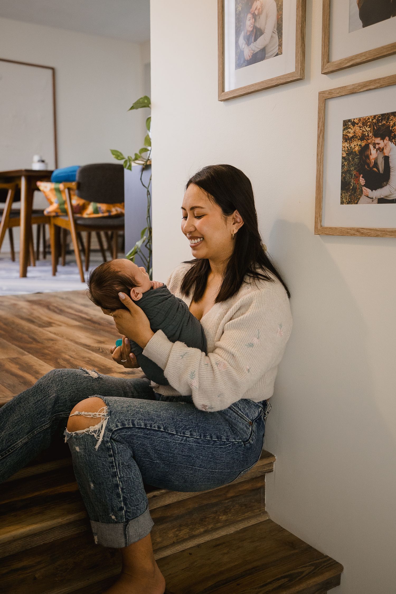 A smiling young woman sits on stairs holding a newborn baby in a cozy home with framed photos on the wall.
