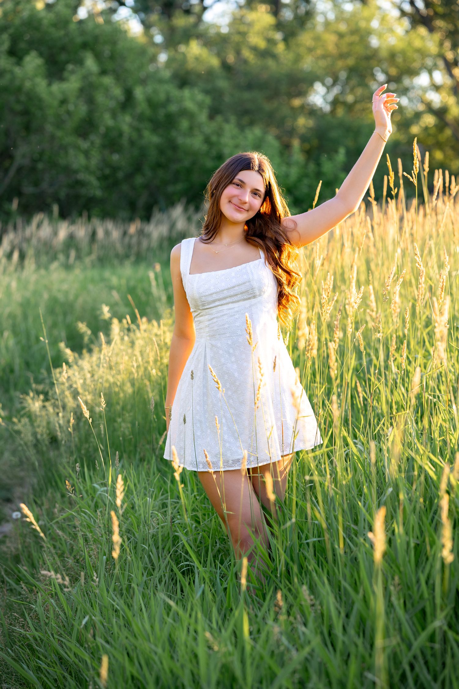Young woman in white dress standing in tall green grass field, raising her hand, bathed in golden sunset light.