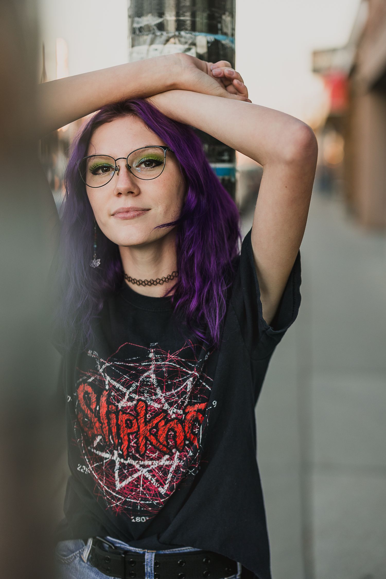 Young woman with purple hair and green glasses wearing a Slipknot band tee, posing outdoors on a city street.