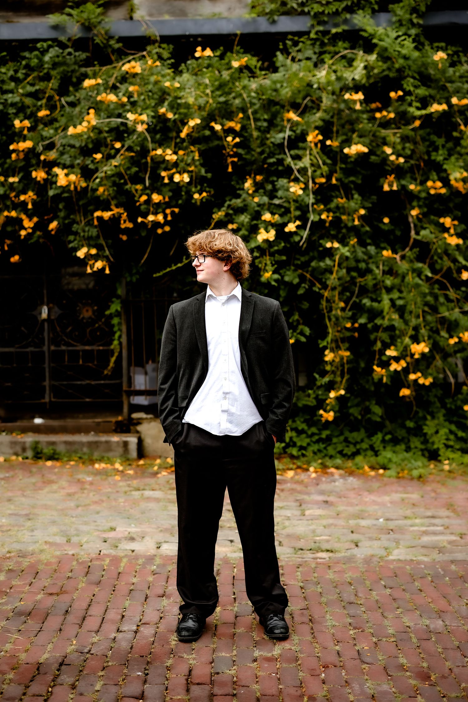 Young man in black suit stands on brick courtyard before yellow flowering vine wall, looking aside confidently.