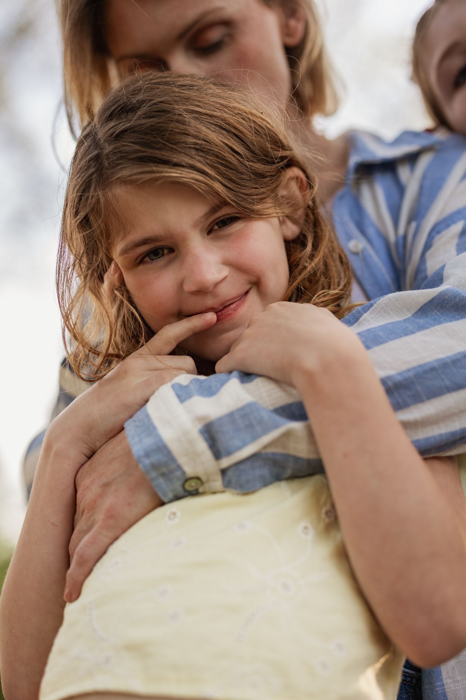 A smiling young girl in a yellow dress being hugged by a woman outdoors in natural light.