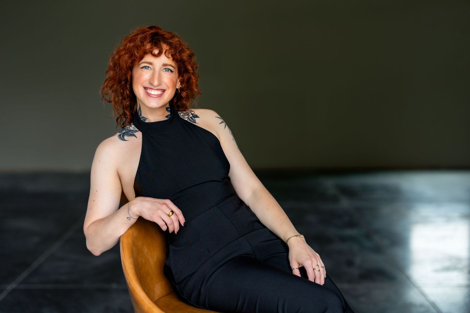 Woman with curly red hair smiling in black halter jumpsuit, seated in amber chair against dark background.