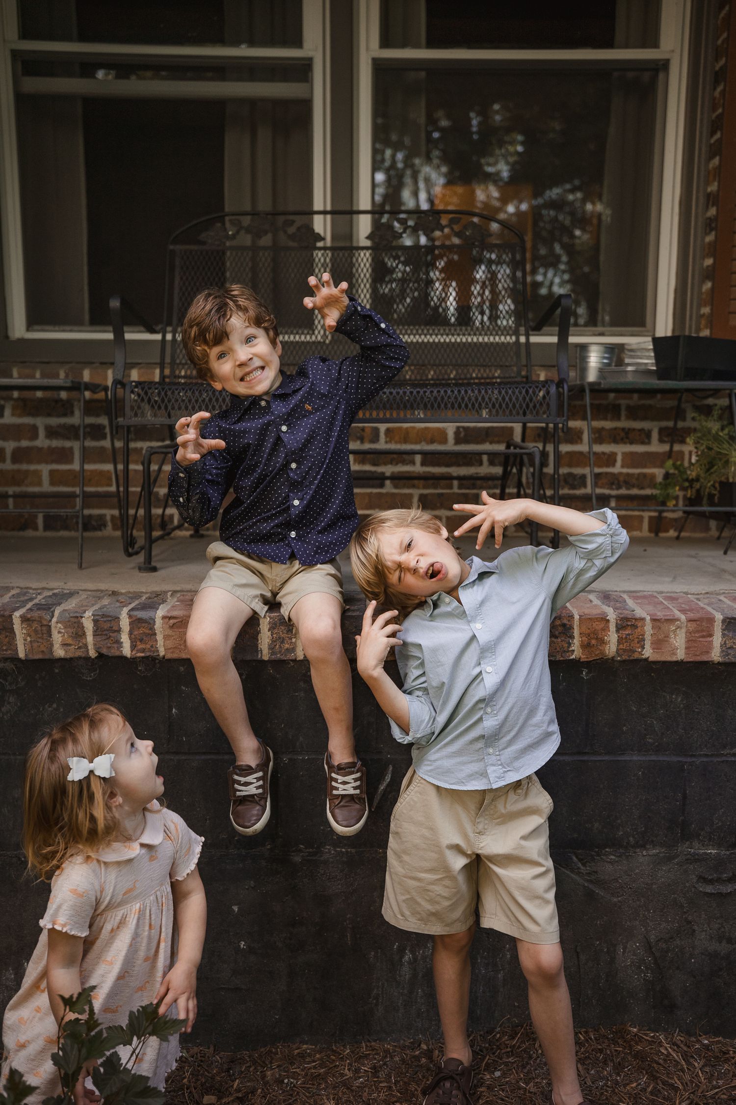 Three playful children pose on brick steps outside a home, two boys making silly faces and a little girl in a white dress.
