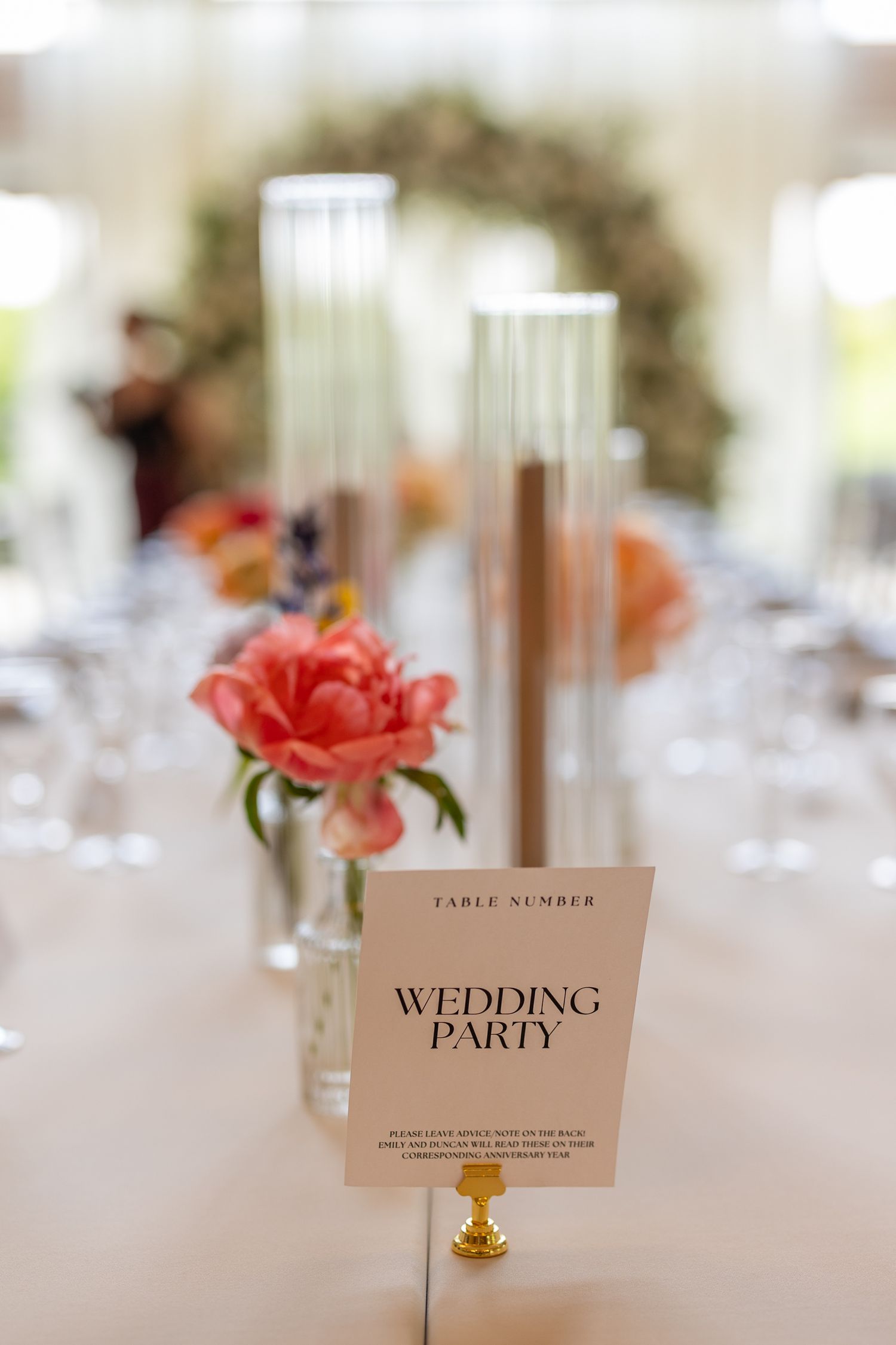 Elegant wedding party table sign with pink peonies, tall glass vases, and crystal glasses at a reception.