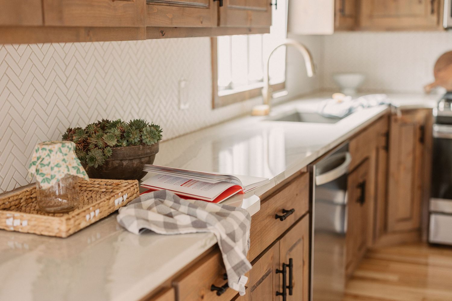 Warm farmhouse kitchen with wood cabinets, white quartz countertops, herringbone tile backsplash, and succulents.