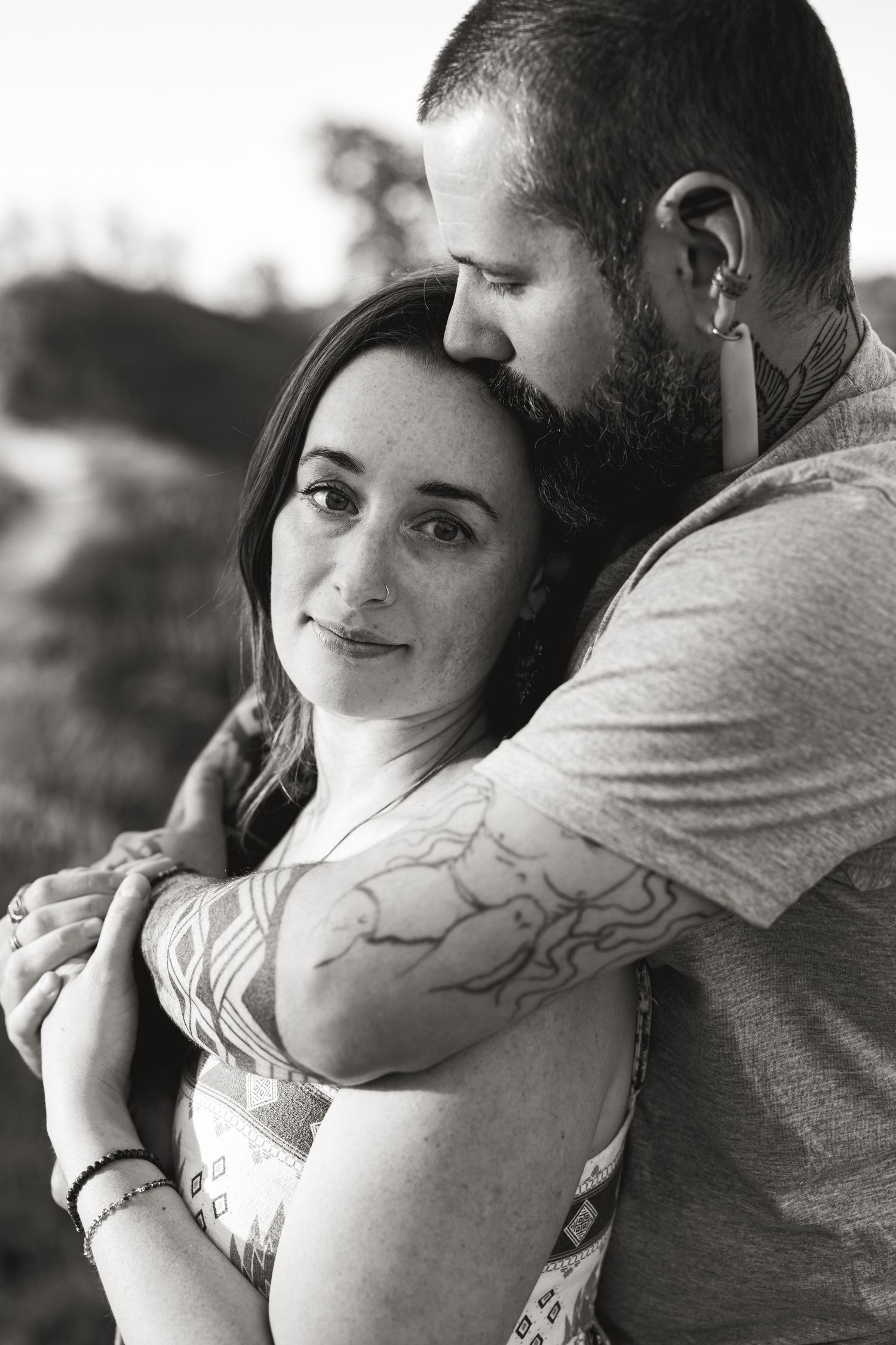 Black and white photo of a bearded man with tattoos embracing a woman with freckles outdoors.