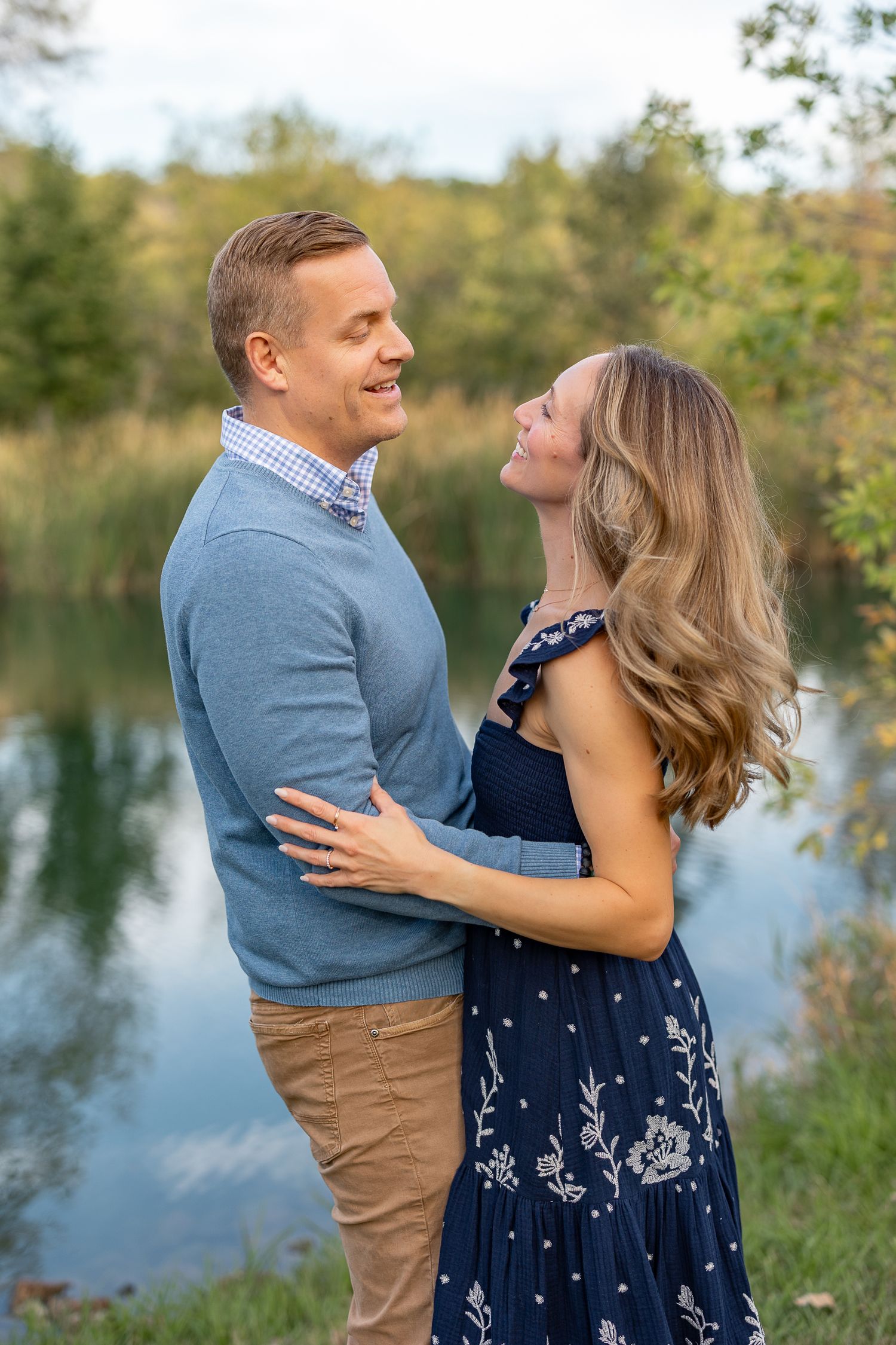 A smiling couple embraces near a reflective pond, the woman in a navy floral dress and man in a blue sweater.