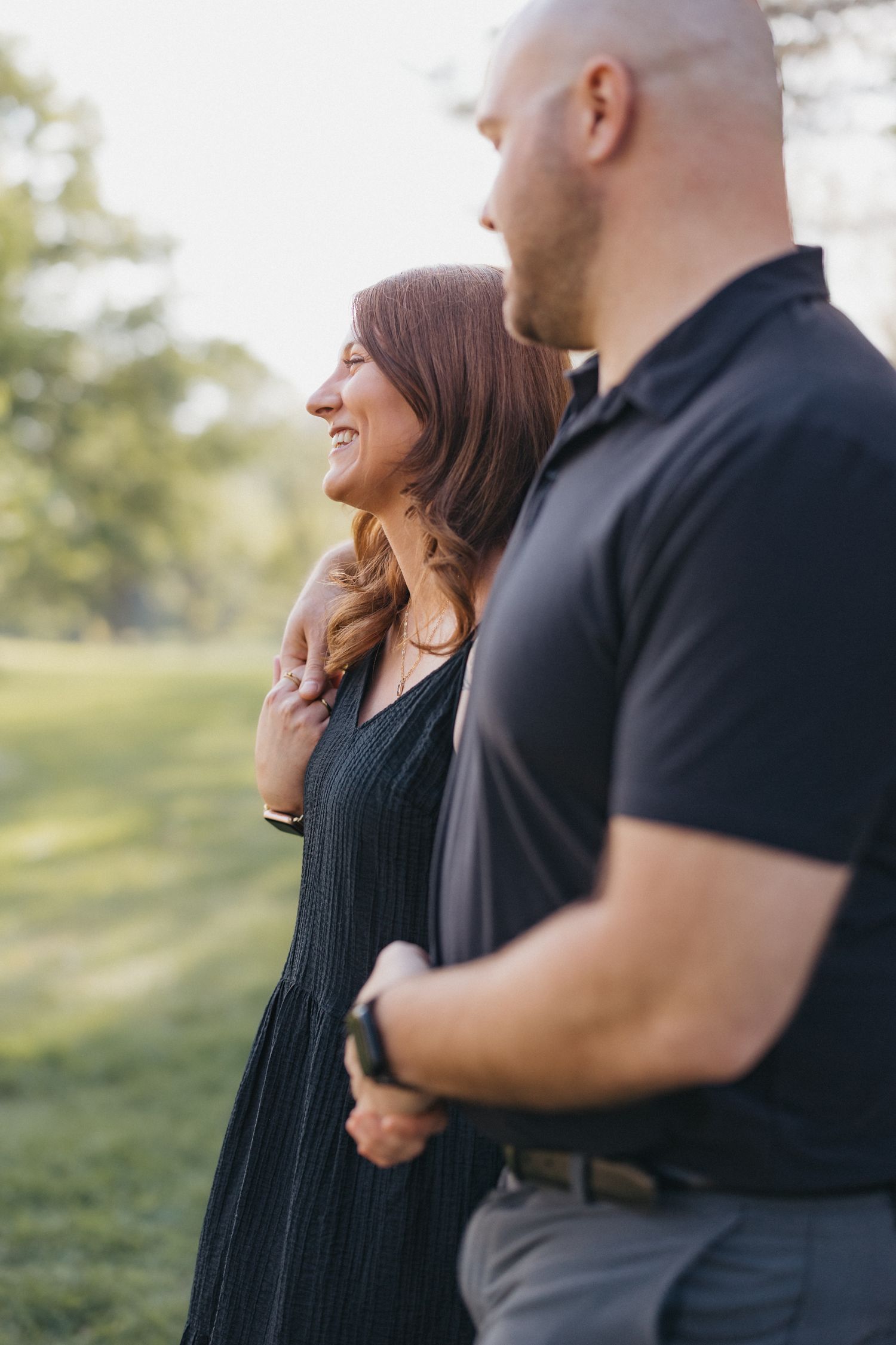 A couple stands together outdoors, the woman smiling in a black dress as the man places his hand on her shoulder.