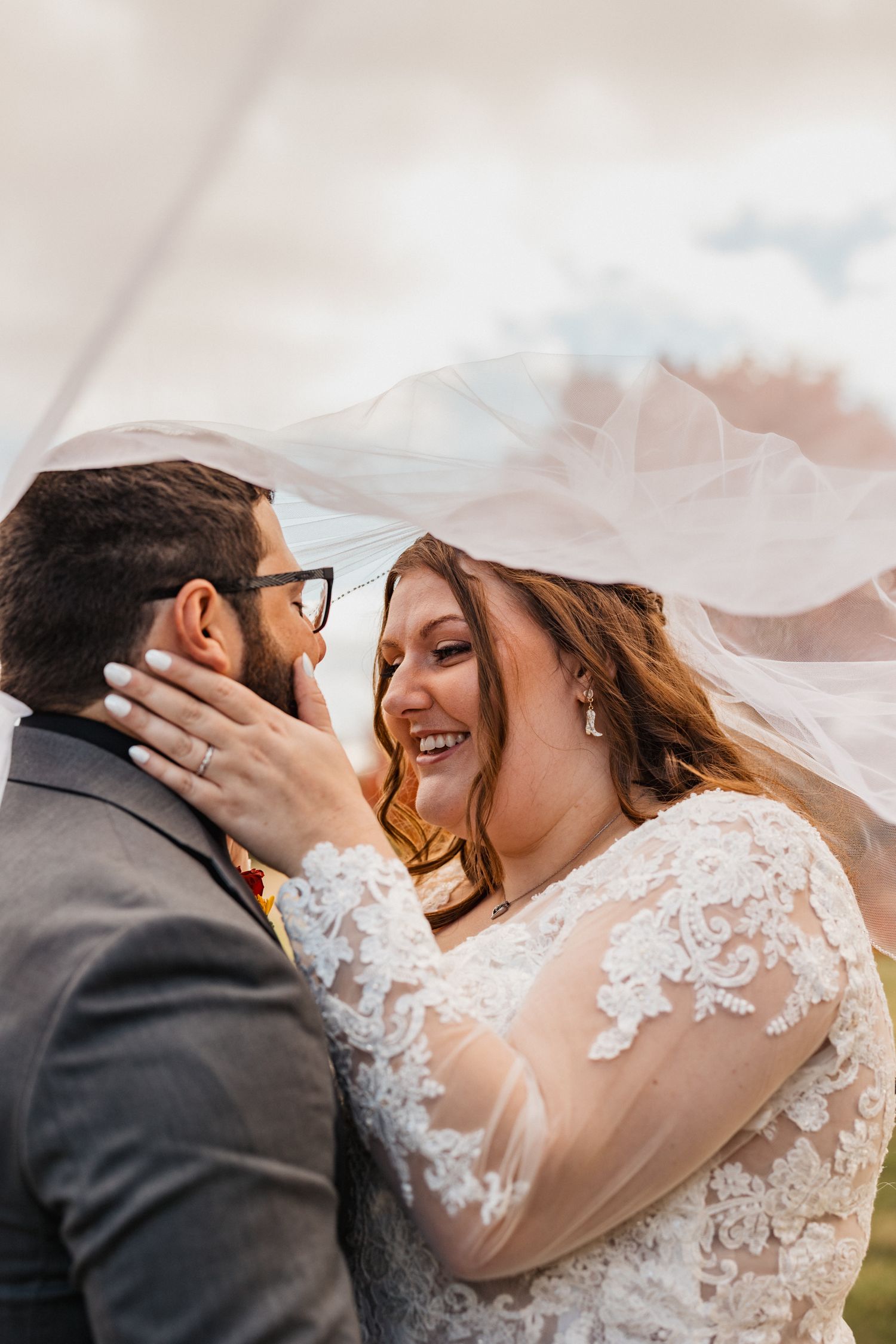 A joyful bride in lace gown laughs with groom under flowing veil at outdoor wedding ceremony.