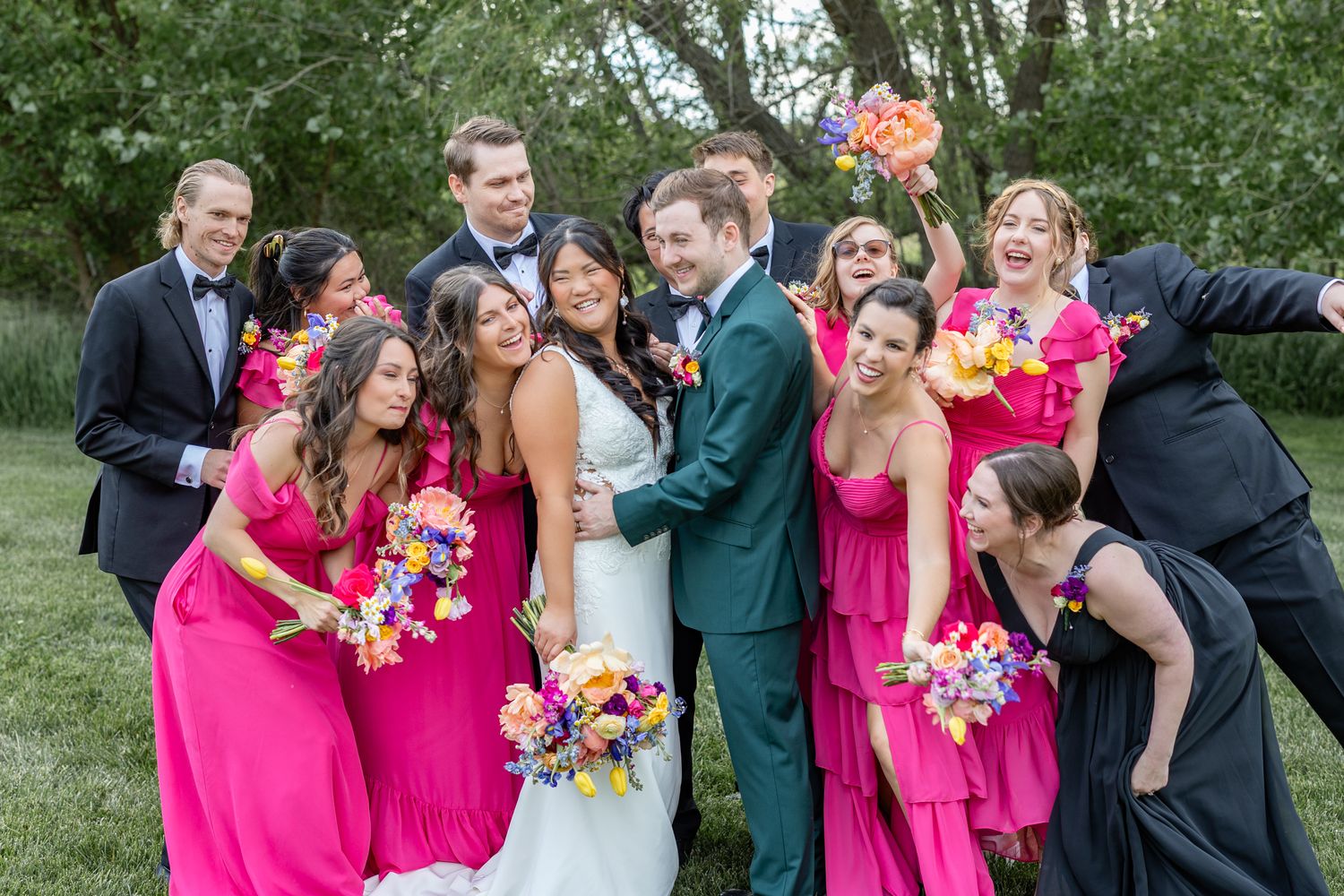 Wedding party in pink bridesmaid dresses and dark suits laugh together outdoors, bride and groom centered.