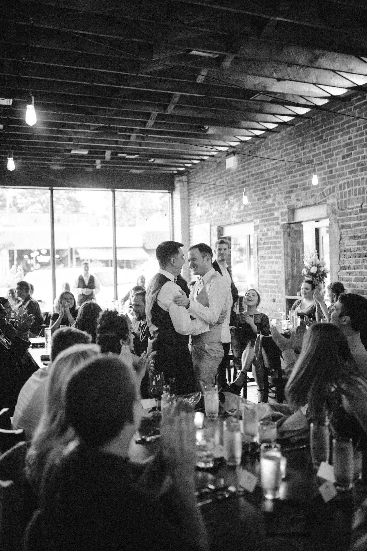 Black and white photo of two grooms sharing a first dance at their wedding reception in a rustic brick venue.
