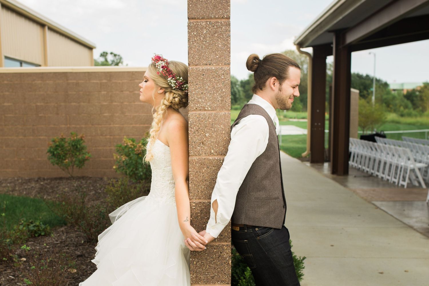 Bride and groom hold hands around a brick pillar on their wedding day, bride wearing floral crown and white gown.