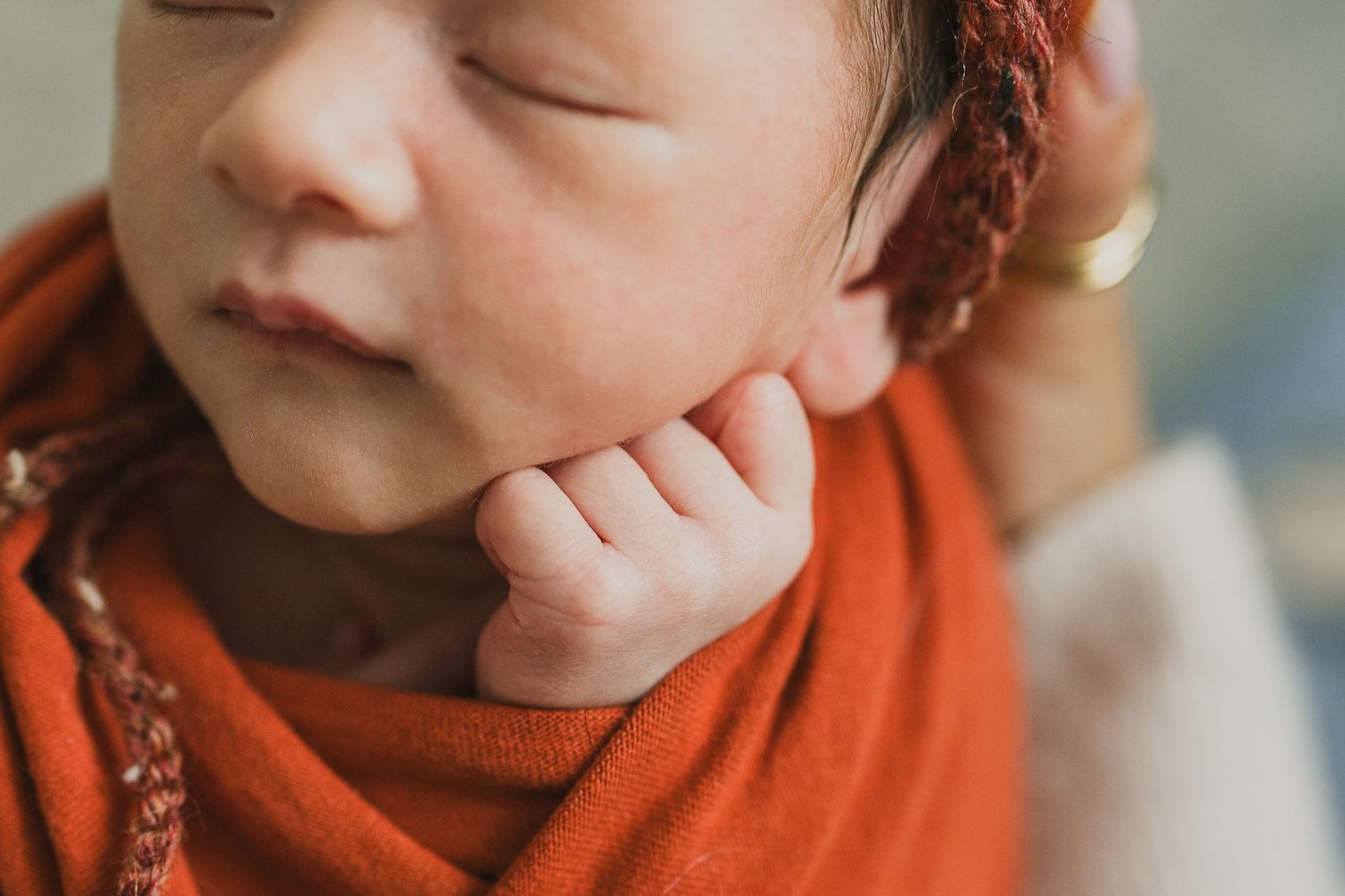 Peaceful newborn baby wrapped in orange swaddle, eyes closed, tiny hand resting on cheek, wearing knit hat.