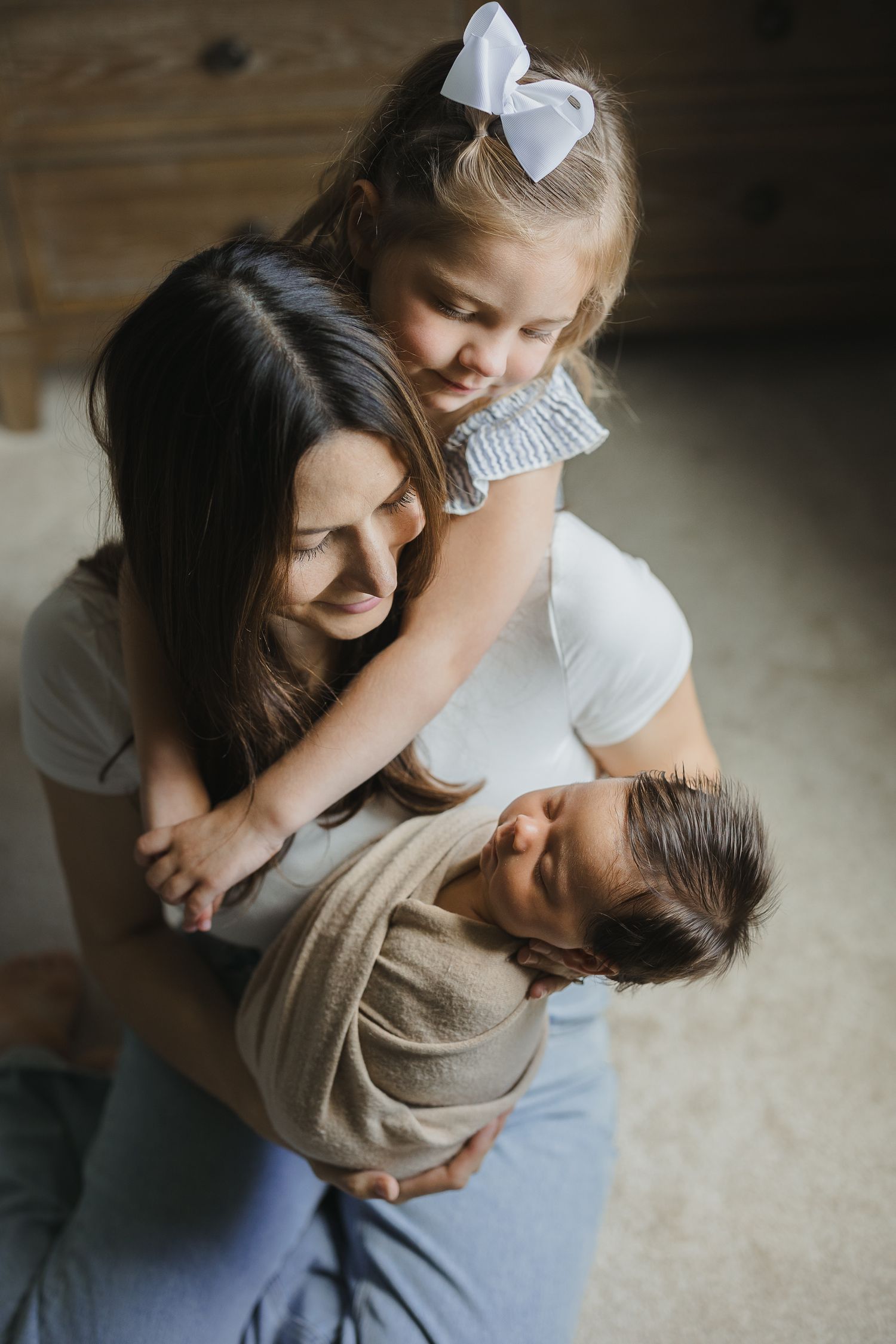 A mother holds her swaddled newborn while her young daughter with a white bow hugs her from behind indoors.