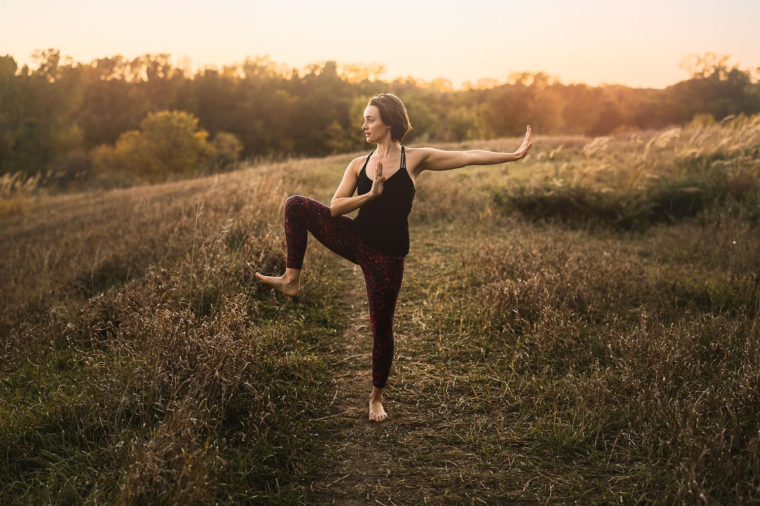 Woman practicing yoga balance pose in a sunlit field at golden hour, wearing dark athletic wear outdoors.