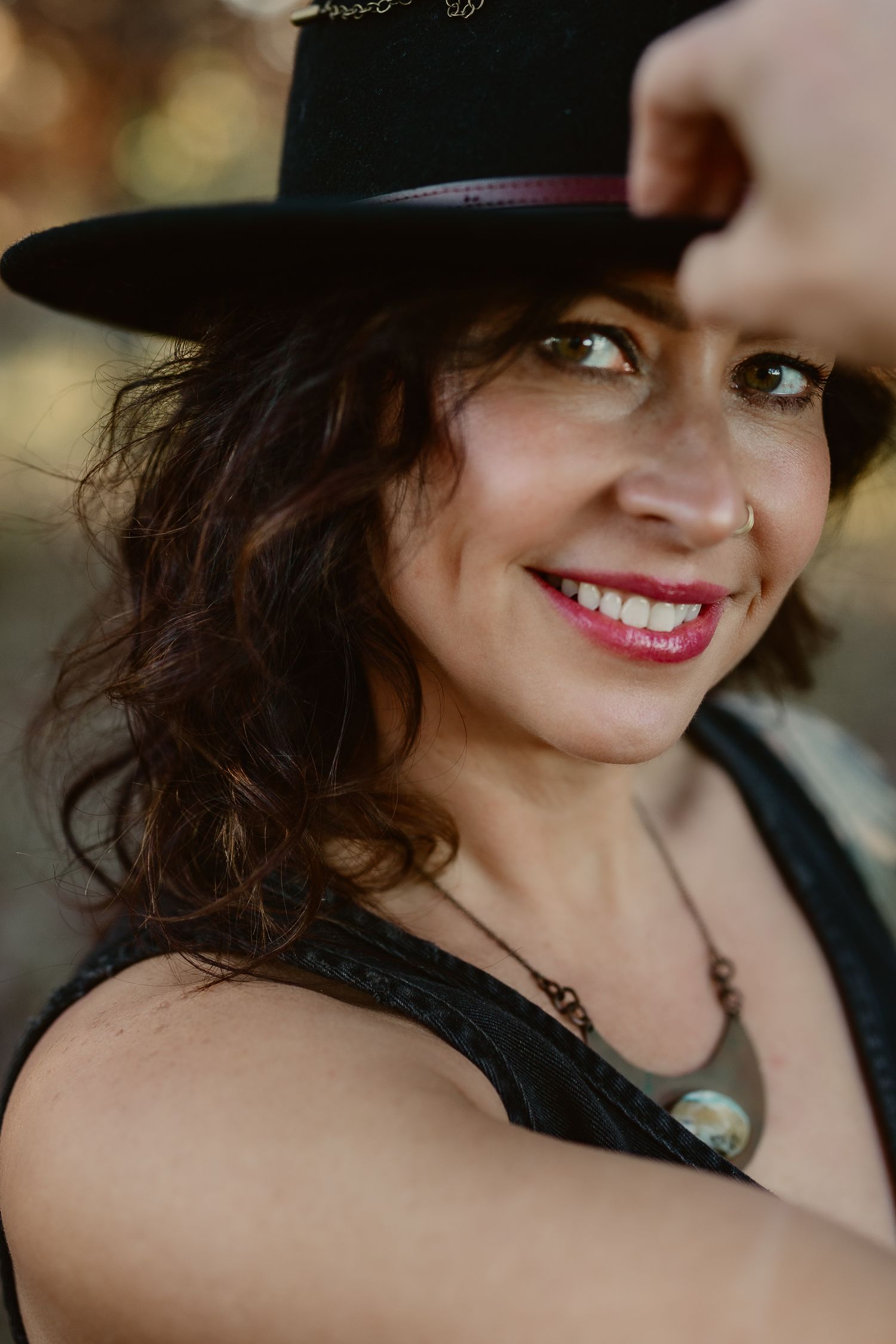 Smiling woman with curly brown hair wearing a black wide-brim hat, pink lipstick, and a necklace, outdoors.