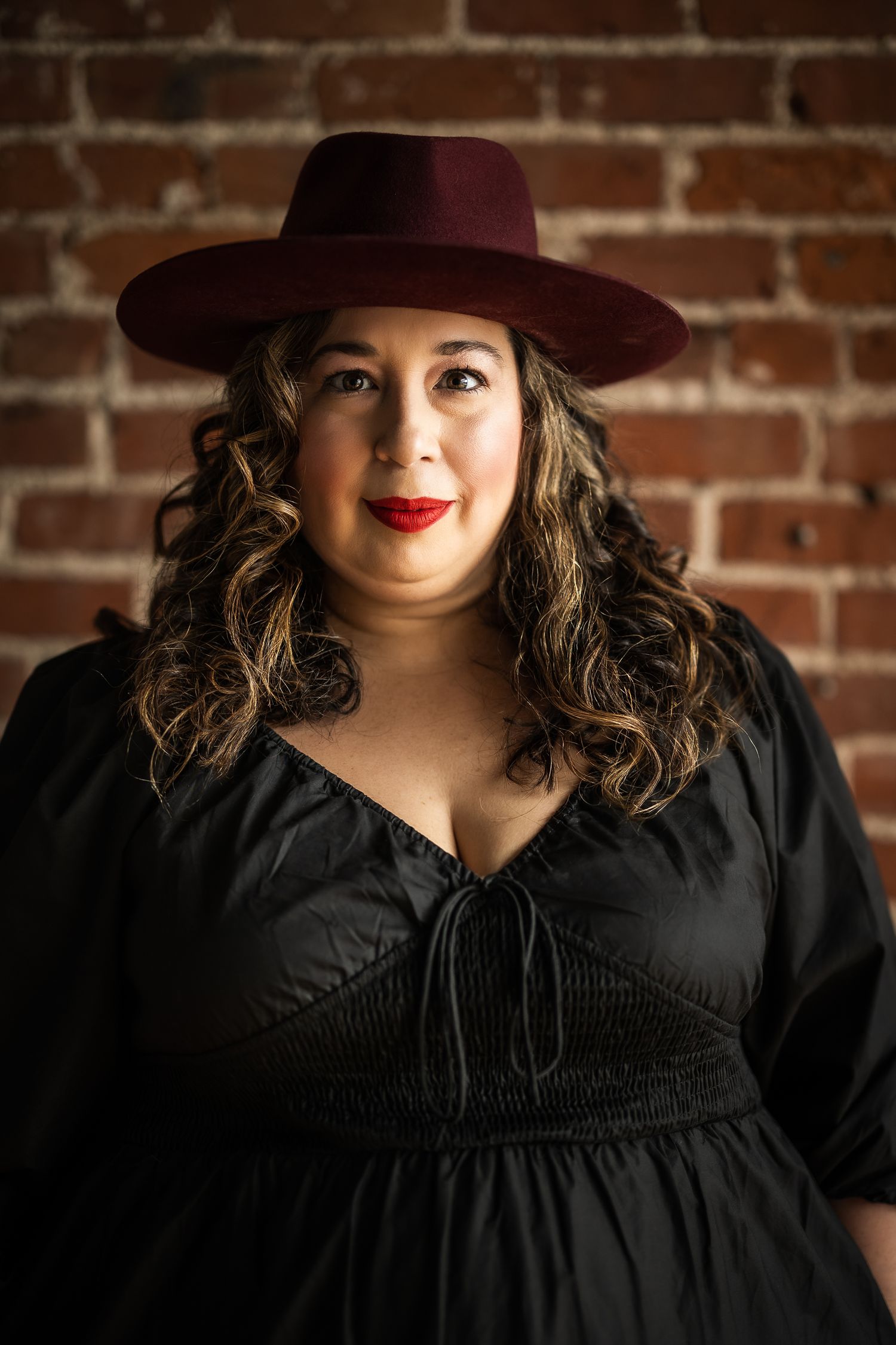 A woman in a black dress and burgundy wide-brim hat poses confidently against a rustic brick wall.