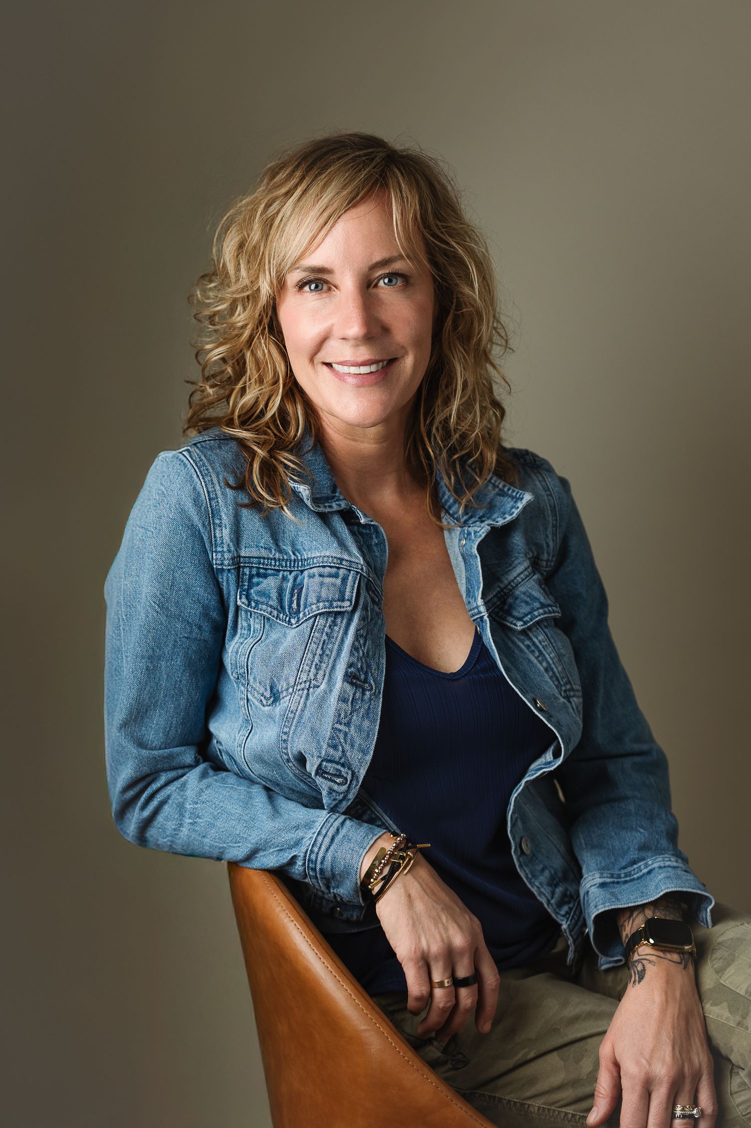 Smiling woman with curly blonde hair wearing a denim jacket and navy top, seated casually in a leather chair.