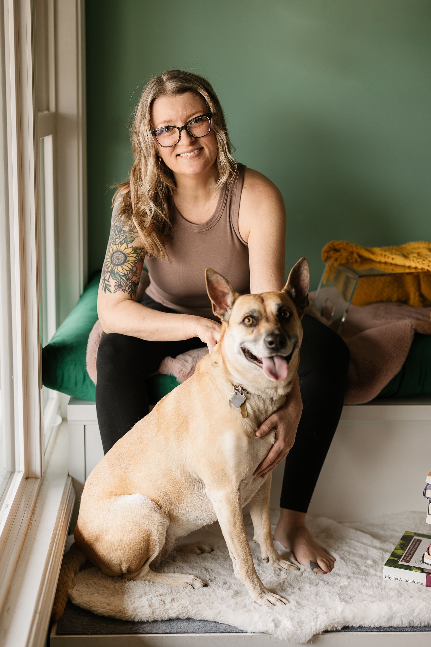 Smiling woman with glasses and tattoos poses with her happy tan dog on a white rug by a green wall.