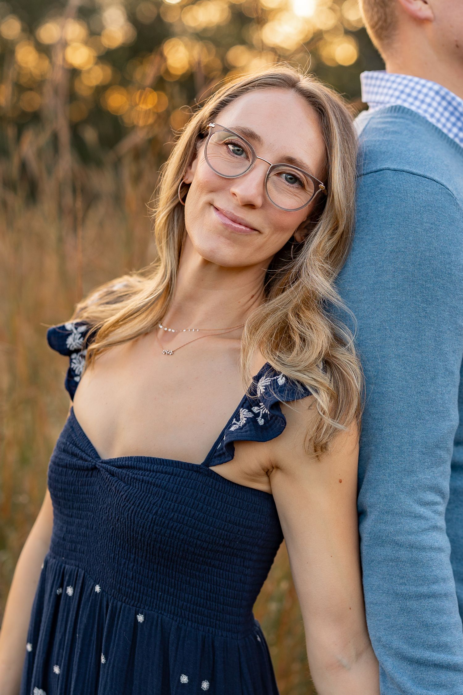 A smiling blonde woman with glasses in a navy dress leans against a man outdoors in golden hour light.
