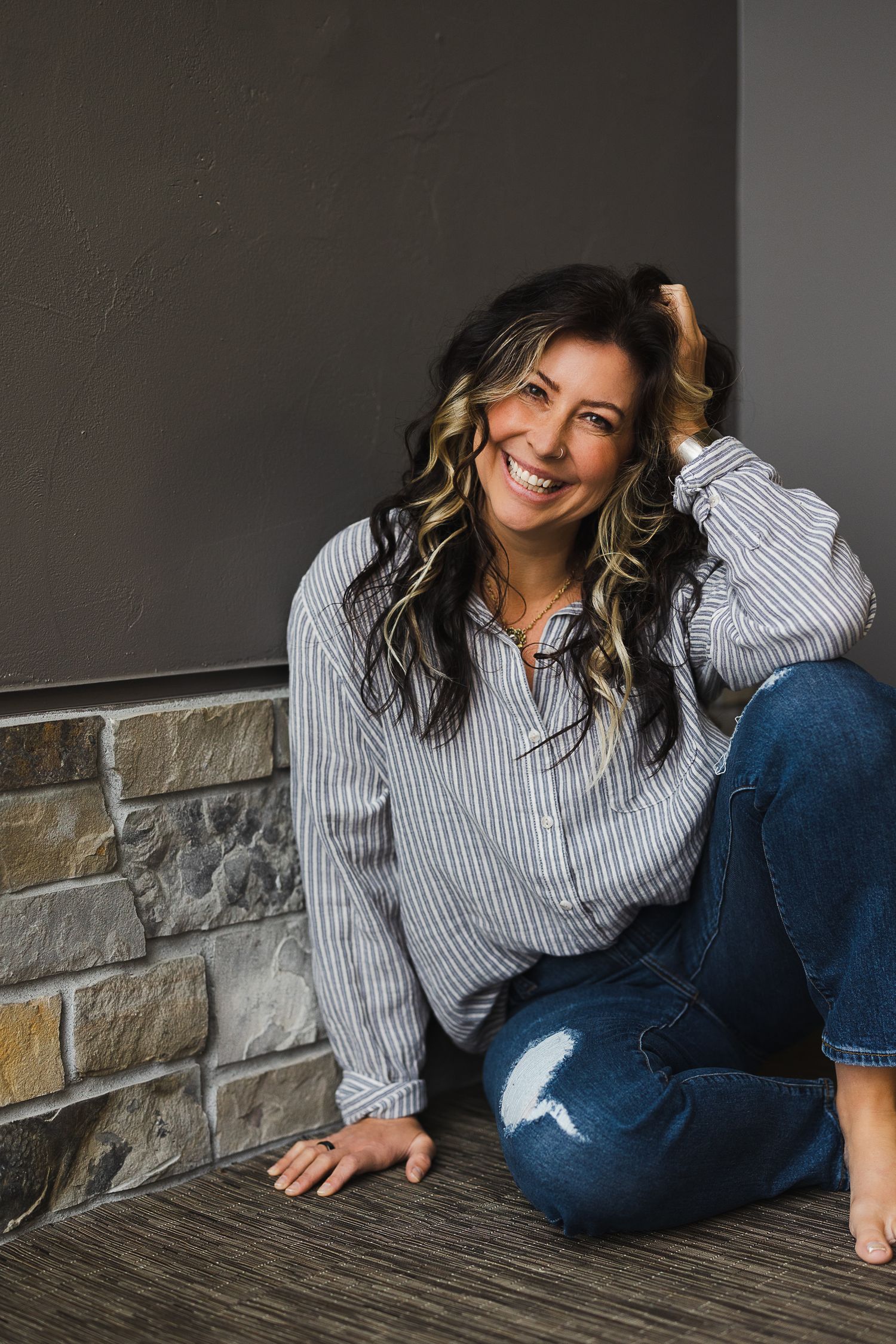 Smiling woman with dark wavy hair sitting on floor in striped shirt and ripped jeans near stone wall.