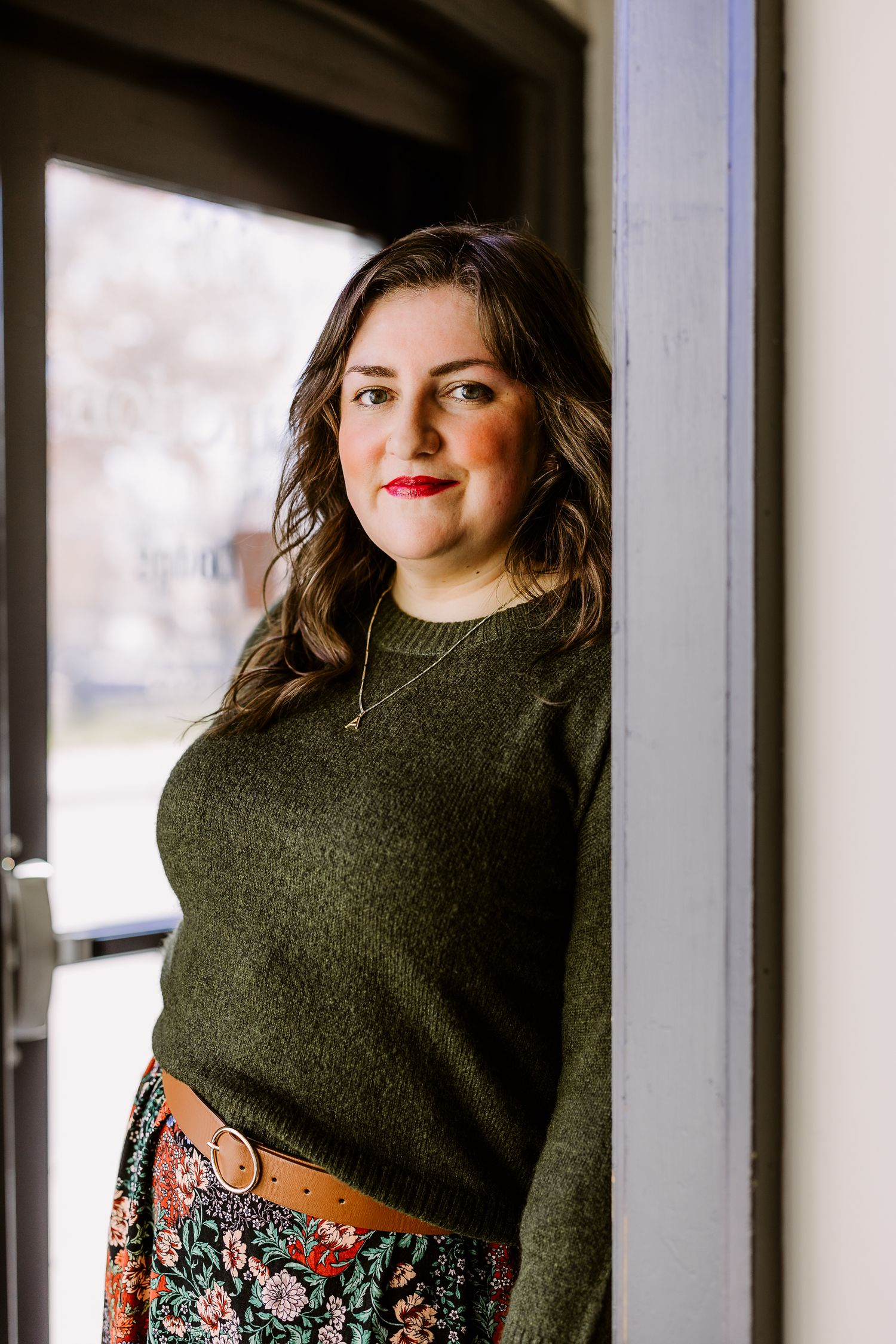 Woman in olive green sweater and floral skirt with brown belt poses by a doorway, wearing red lipstick.