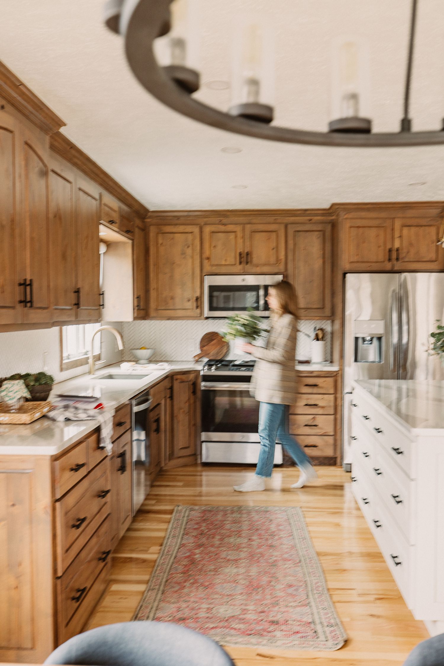 Woman walking through a rustic kitchen with warm wood cabinets, stainless steel appliances, and a vintage red runner rug.