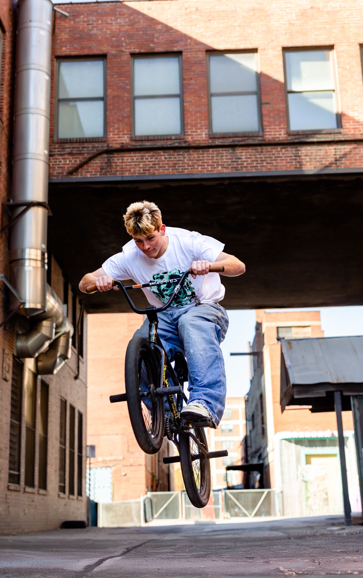 Young BMX rider performs an aerial trick on his bike in an urban alleyway between brick buildings.