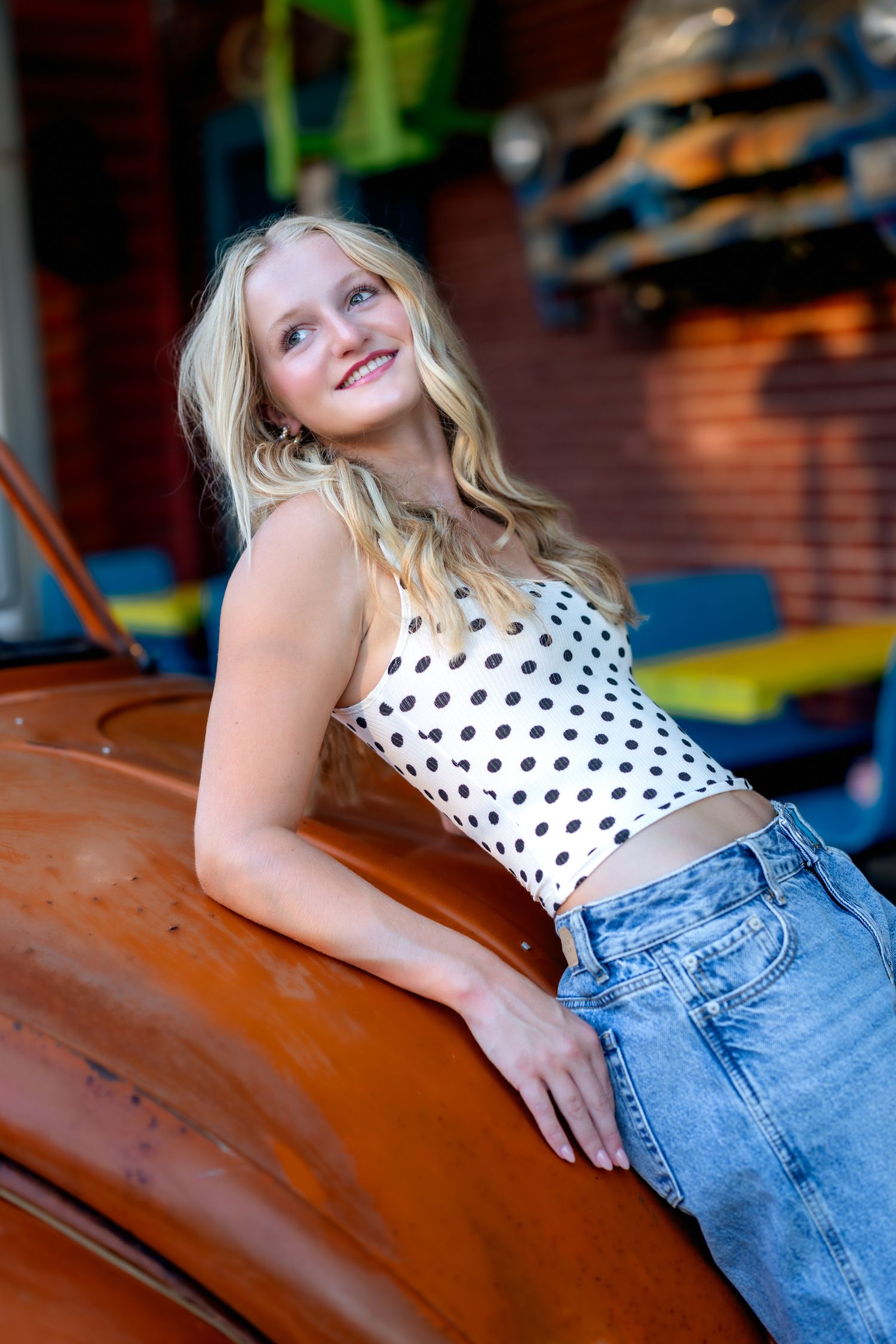 Smiling blonde woman in polka dot crop top and jeans leaning on a vintage car outdoors.