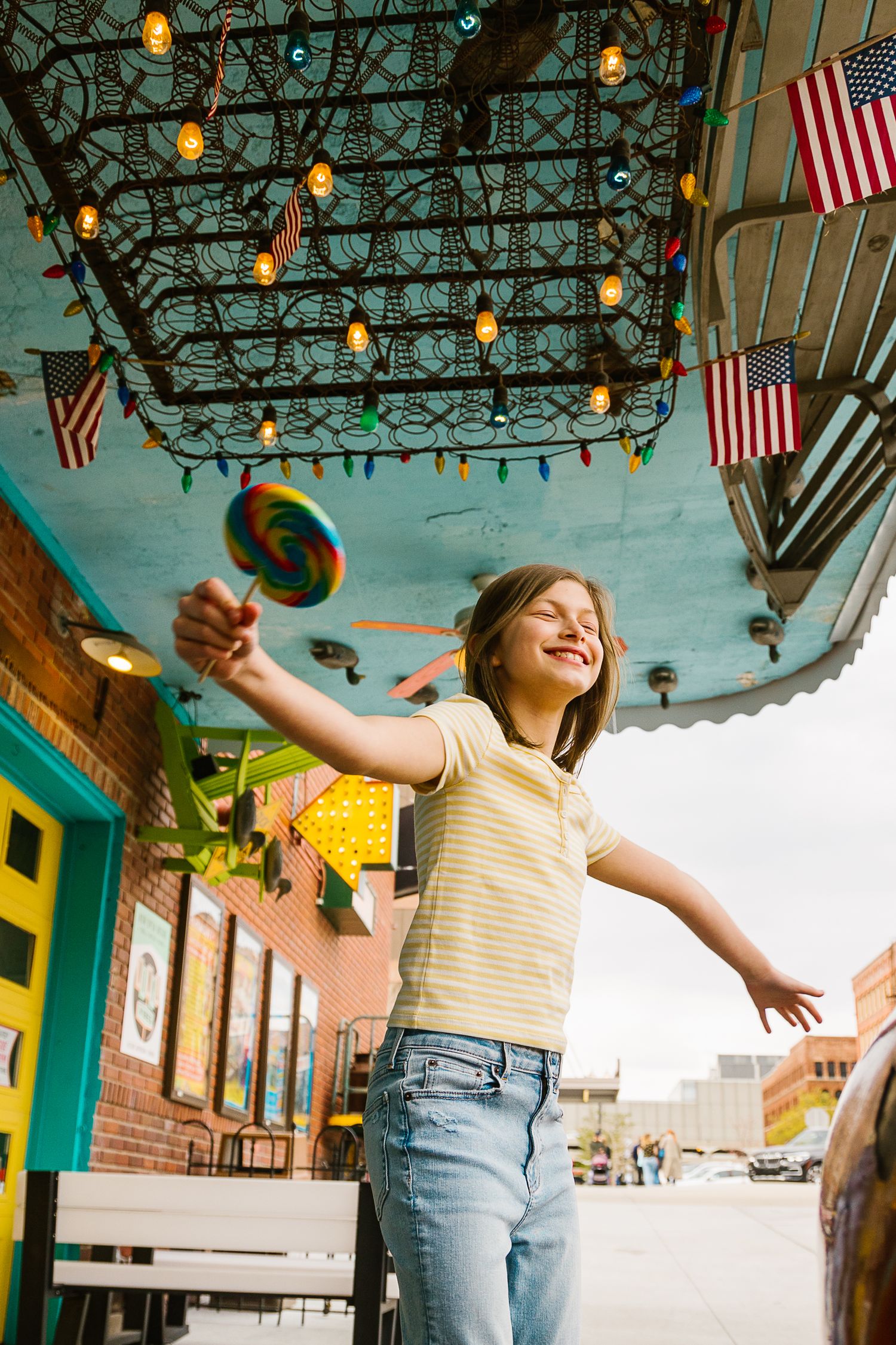 Happy girl holding a colorful lollipop outdoors under string lights and American flags.