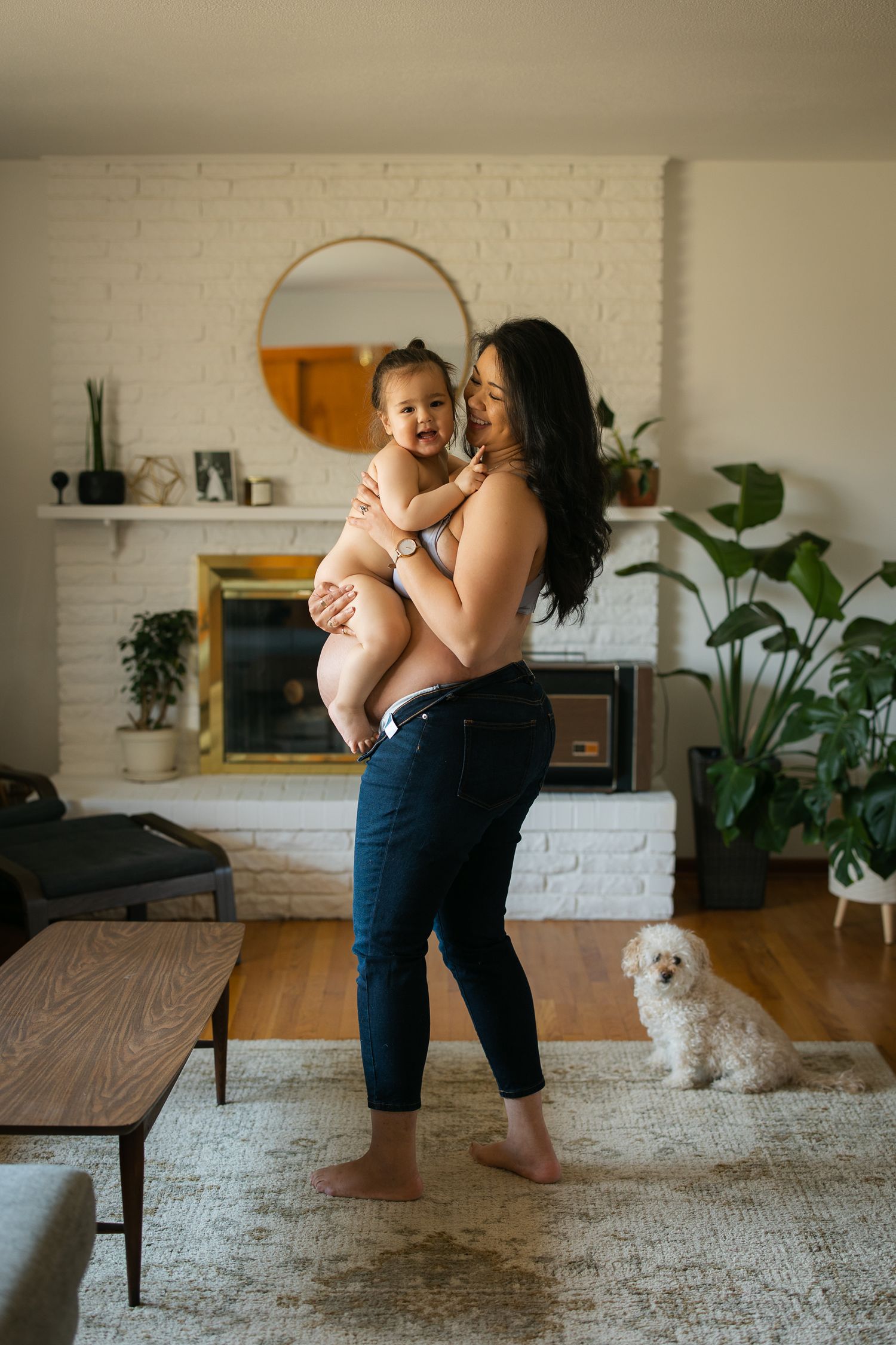 A woman holds a smiling baby in a cozy living room with a fireplace, white brick wall, and a small white dog nearby.