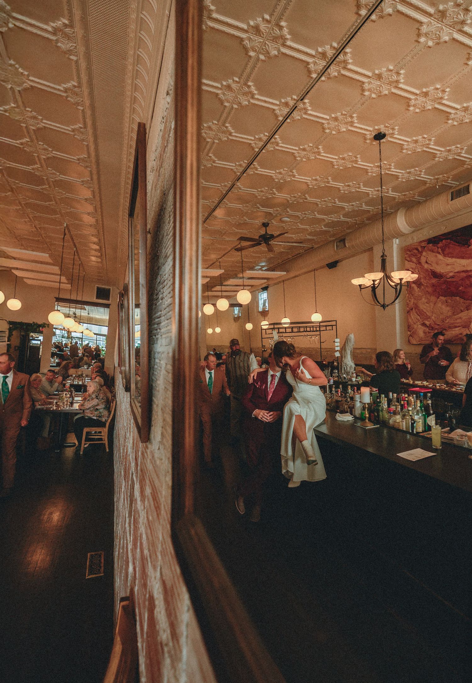 Elegant wedding reception in a vintage restaurant with ornate tin ceilings, warm lighting, and guests celebrating near a bar.