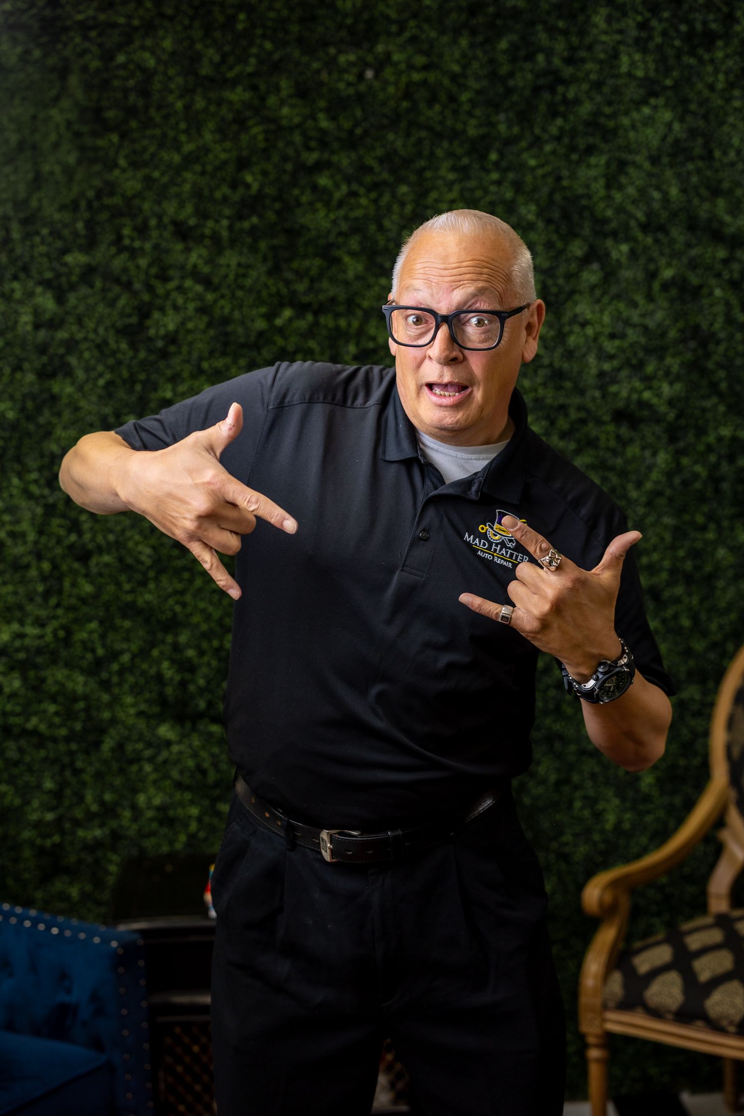 Bald man in black polo shirt with glasses making ASL I love you signs with hands in front of green hedge wall backdrop.