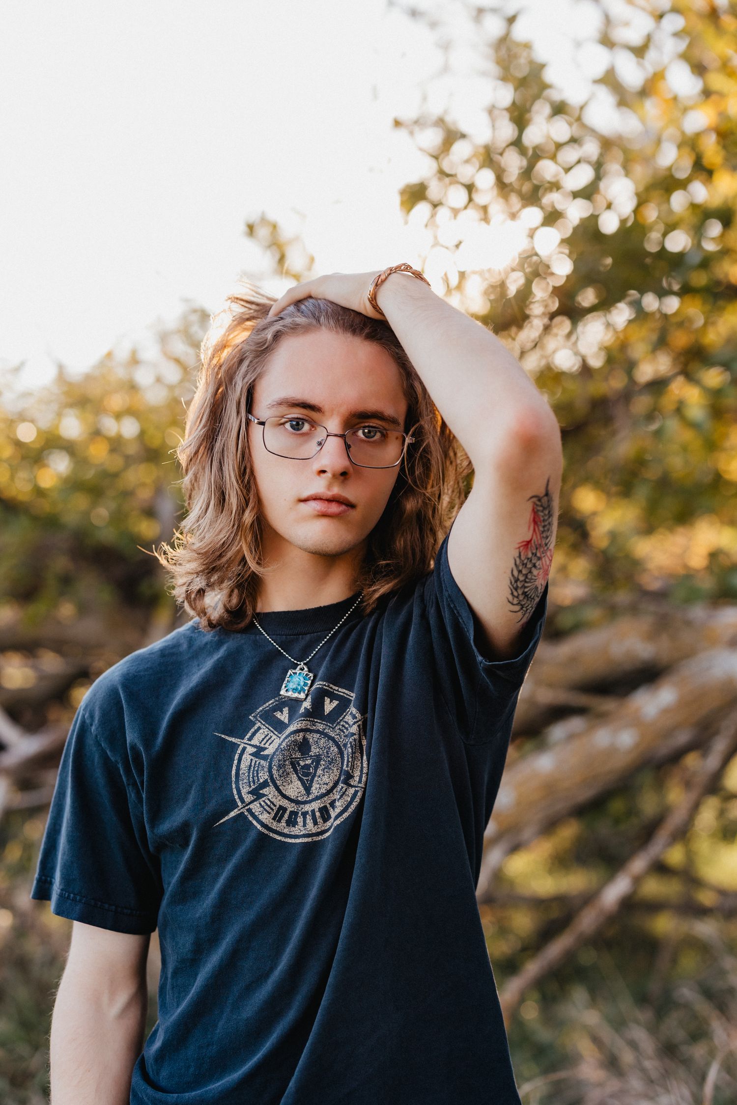 Young man with long hair, glasses, and tattoo wearing a dark graphic tee, posing outdoors in golden hour light.