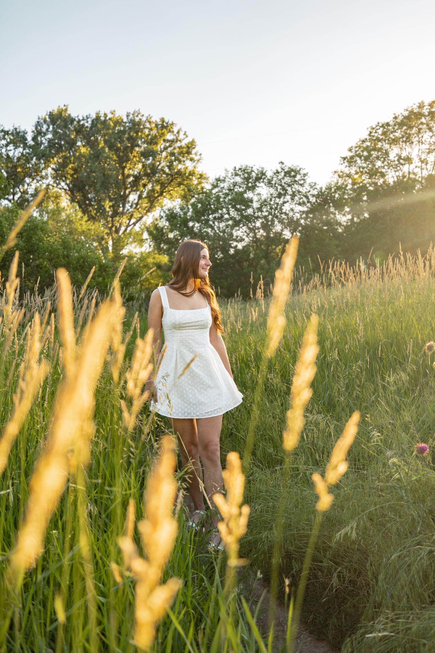 Young woman in white dress standing in sunlit green meadow with tall grass and trees at golden hour.