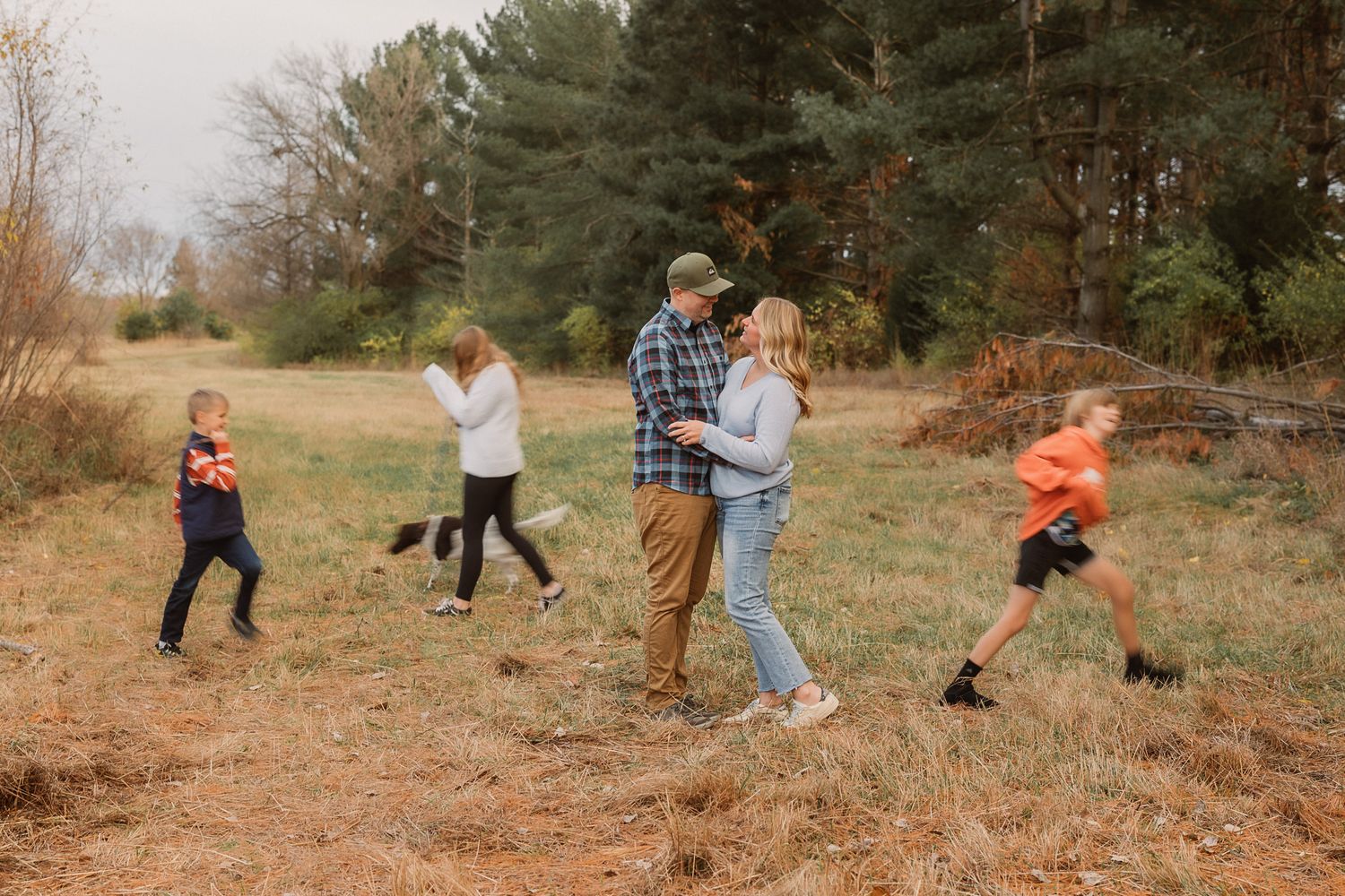 A family poses and plays in an open field surrounded by autumn trees, with children running and parents embracing.