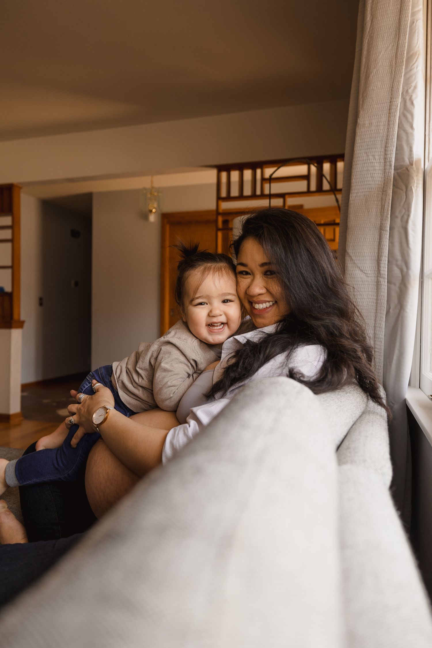 A smiling mother holds her laughing baby on a cozy sofa near a window in a warm, inviting living room.