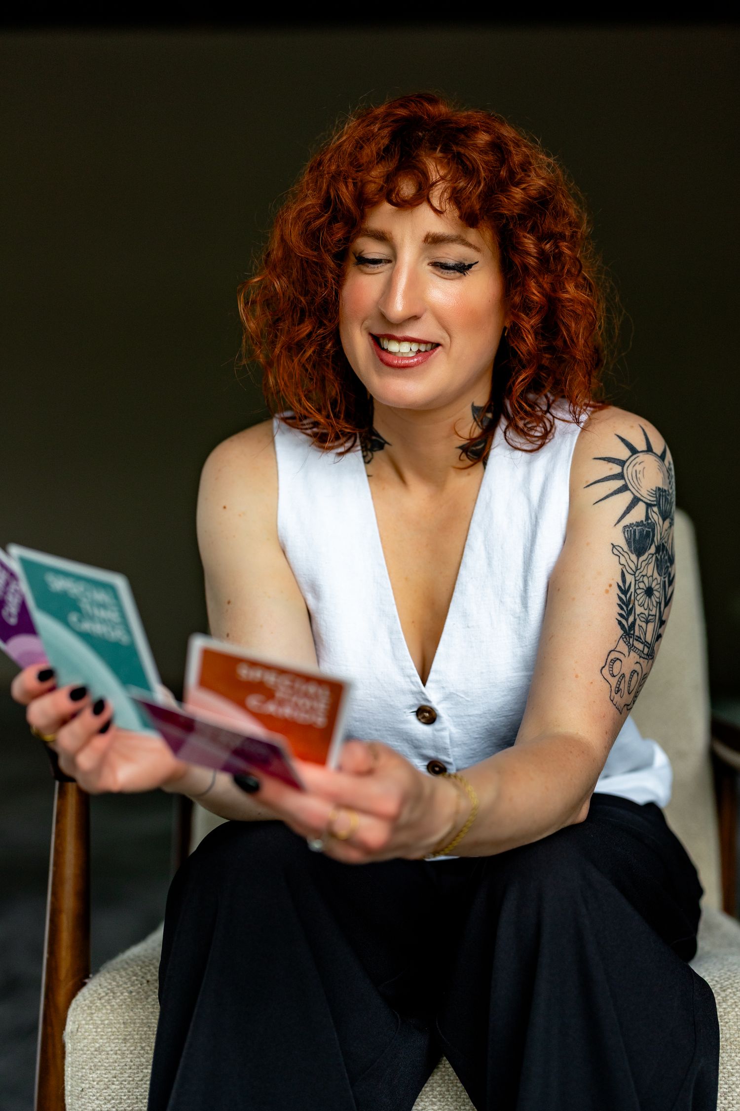 Smiling redhead woman with tattoos reading colorful cards while seated in a white chair.