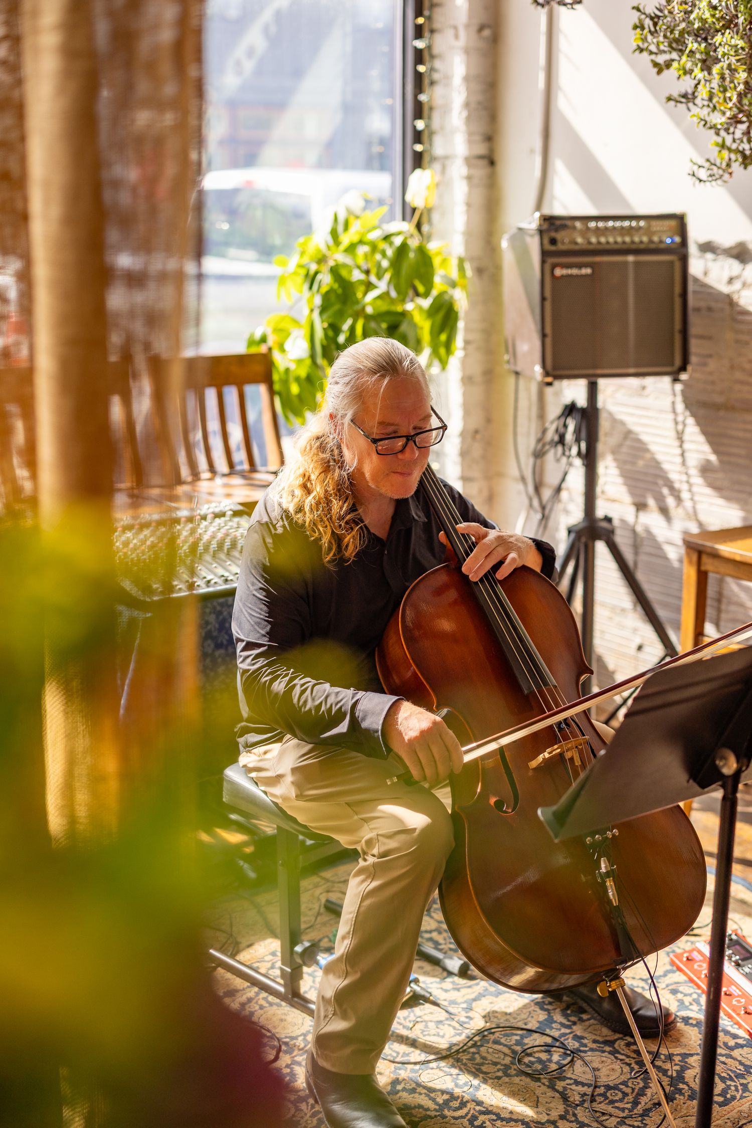 A musician with long gray hair and glasses plays cello indoors, bathed in warm sunlight near a window.