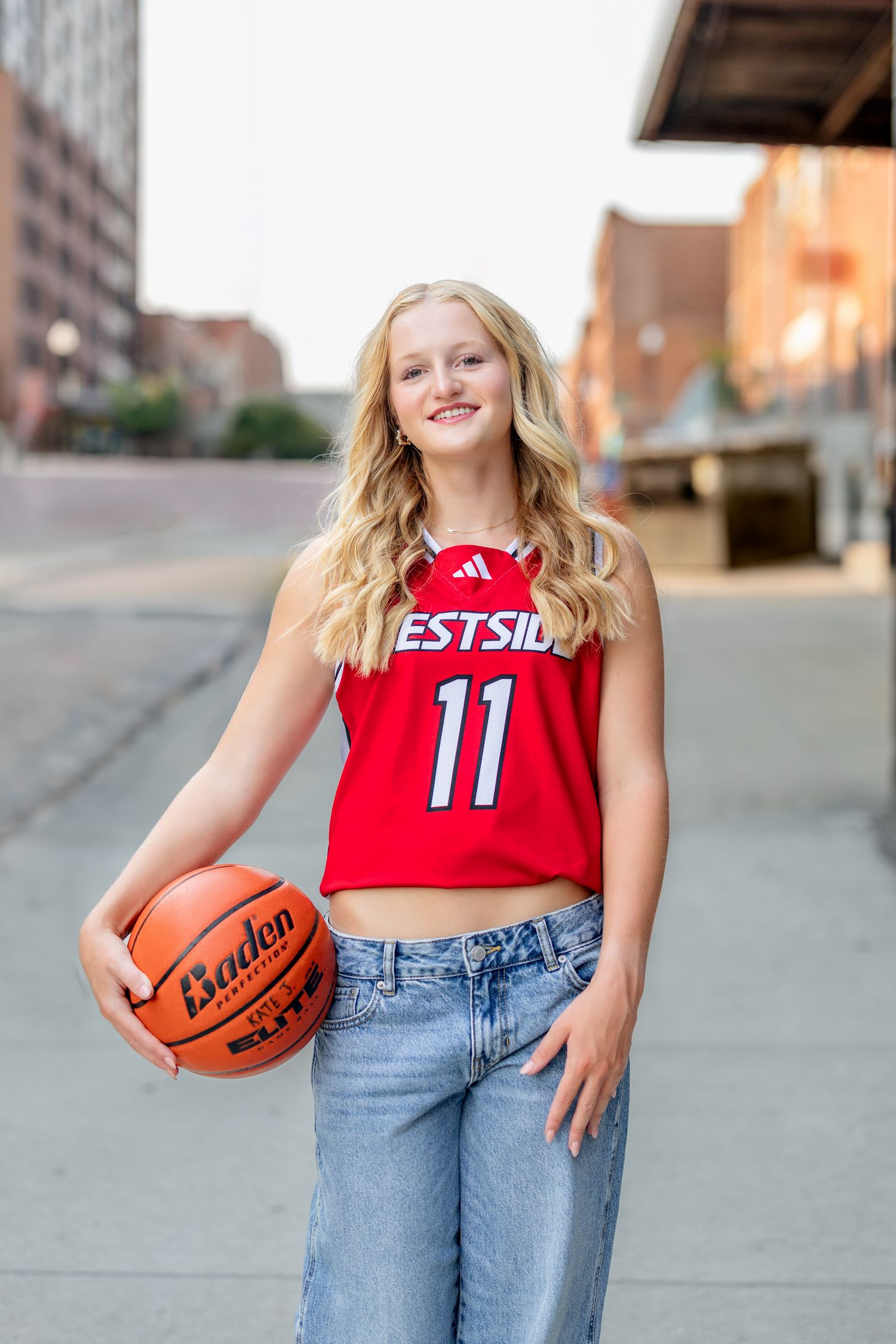 Young blonde girl in red Westside #11 basketball jersey holding a Baden basketball outdoors on a city street.